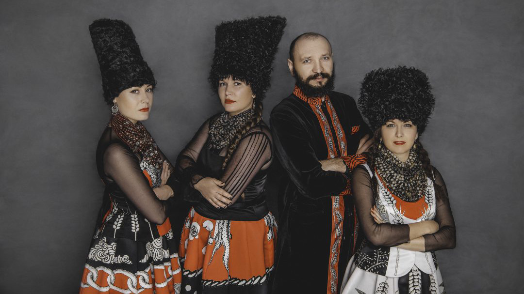 Four people with light complexion stand with their arms crossed for a photo. They are wearing ornate traditional black white and orange Ukrainian outfits. Three have large plush black headwear on