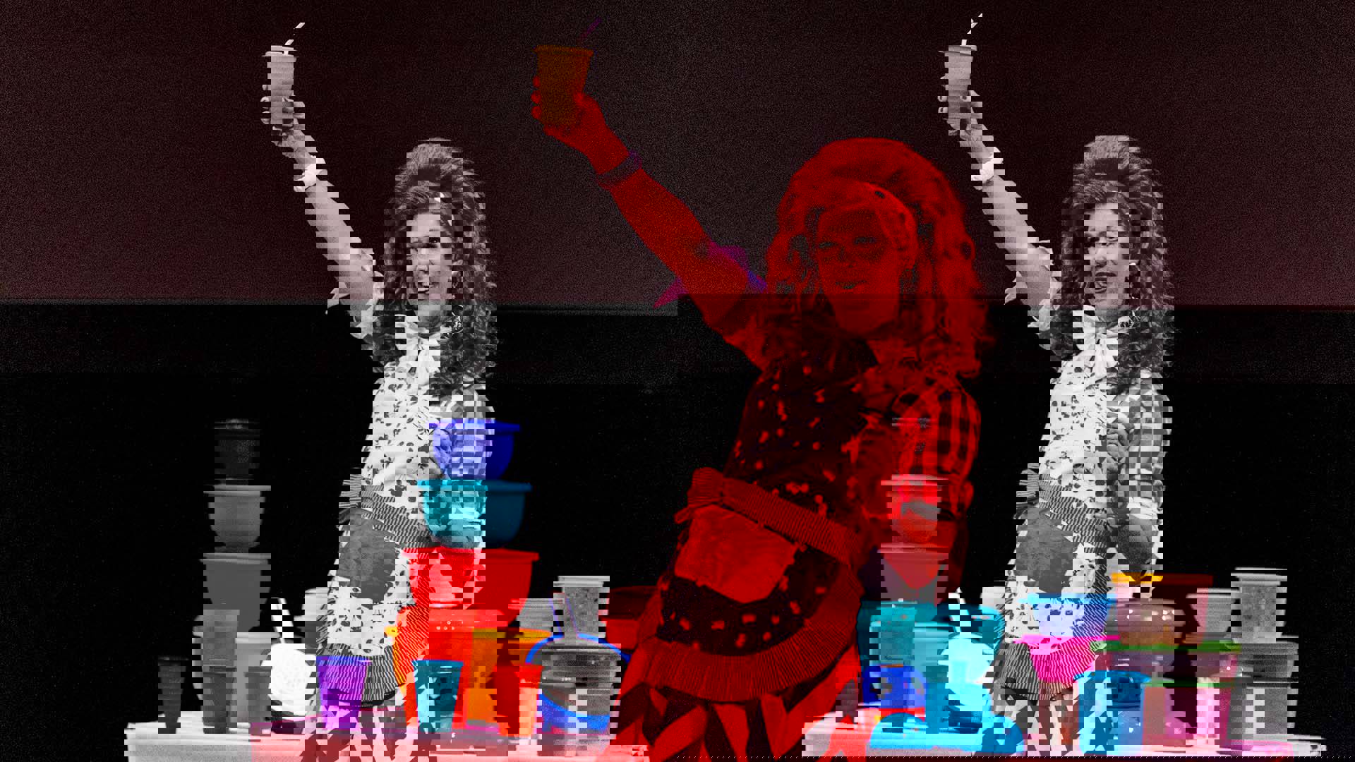 A drag performer with light complexion, voluminous red hair, red lipstick and large diamond drop earrings performing on stage with colorful Tupperware bowls, wearing a red and white gingham dress and cherry print apron. They are raising a yellow cup with a straw with one hand and smiling.