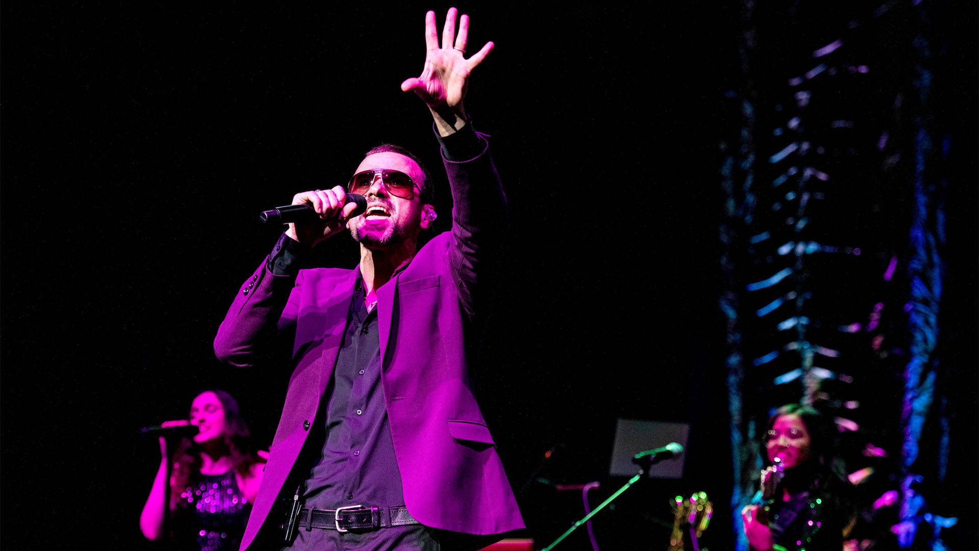 A white man with short brown hair , sunglasses and a purple jacket singing into a microphone with his left hand up in the air.