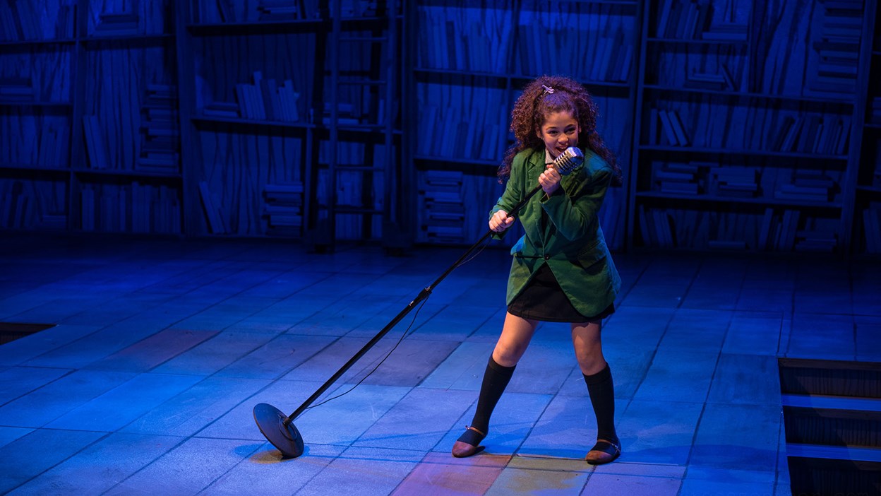 A teenage girl with medium complexion and brown curly hair pulled up is performing on stage, singing into a retro microphone on a stand. She is wearing a forest green blazer and black skirt, long black songs with mary jane flats.