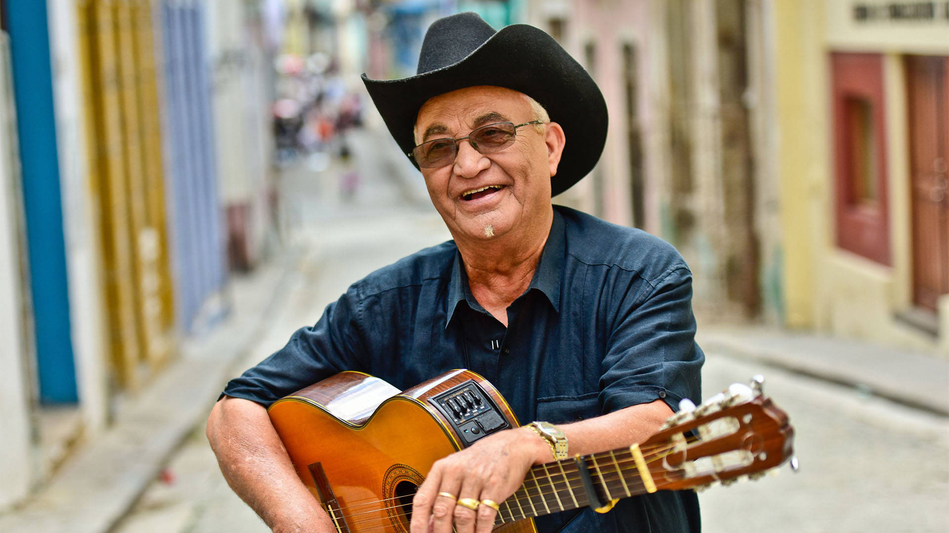 Eliades Ochoa, a Cuban man with medium skin tone and short white hair. He is wearing tinted glasses, a black cowboy hat and a black dress shirt while smiling and holding a guitar.