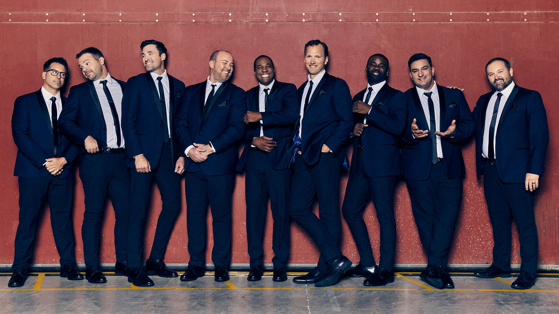 Nine men wearing blue suits standing in front of a red wall. They are smiling and looking in various directions.