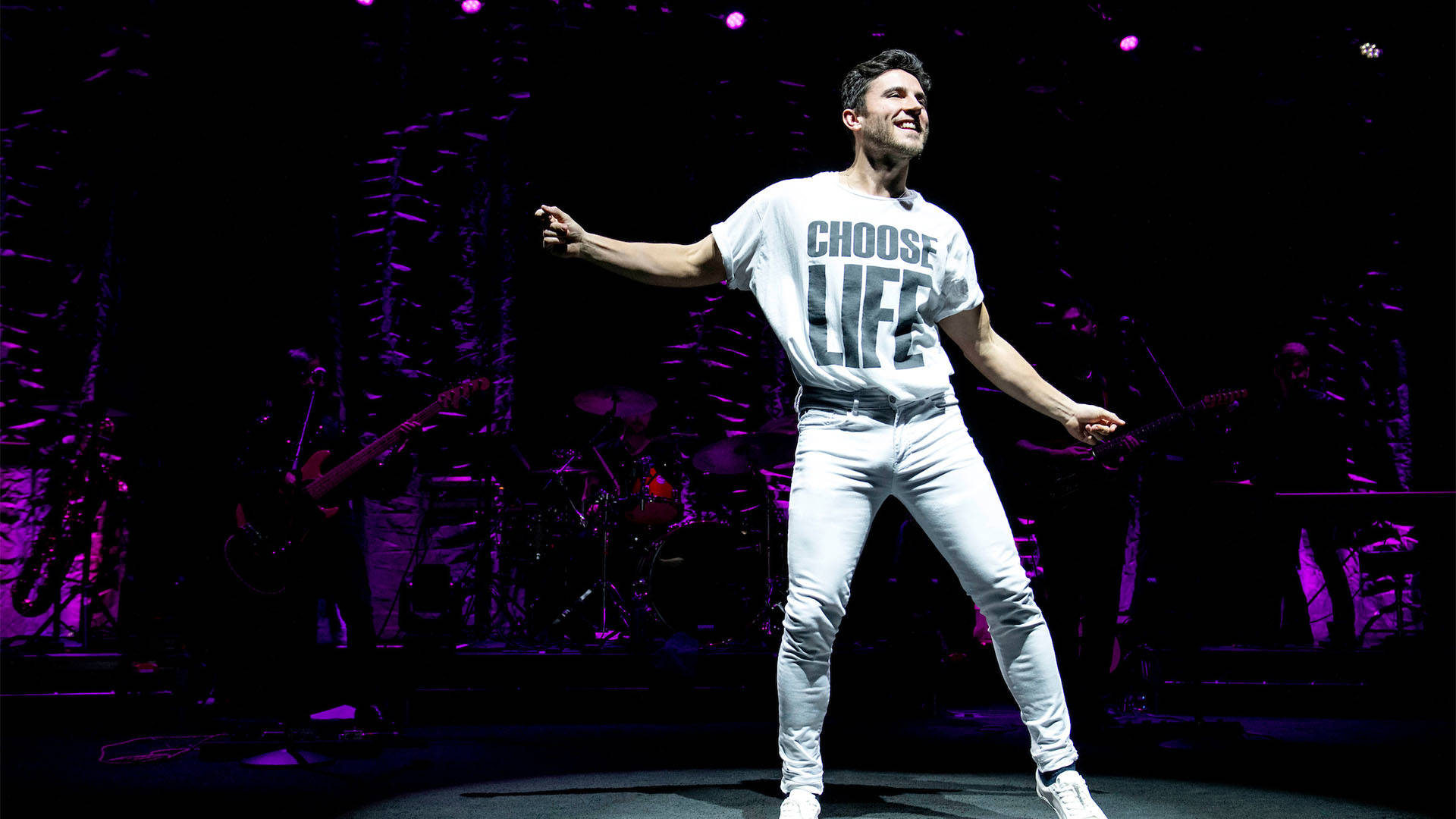 A white man with short brown hair smiles and dances as he performs. He is wearing all white and his shirt has the text "Choose Life"