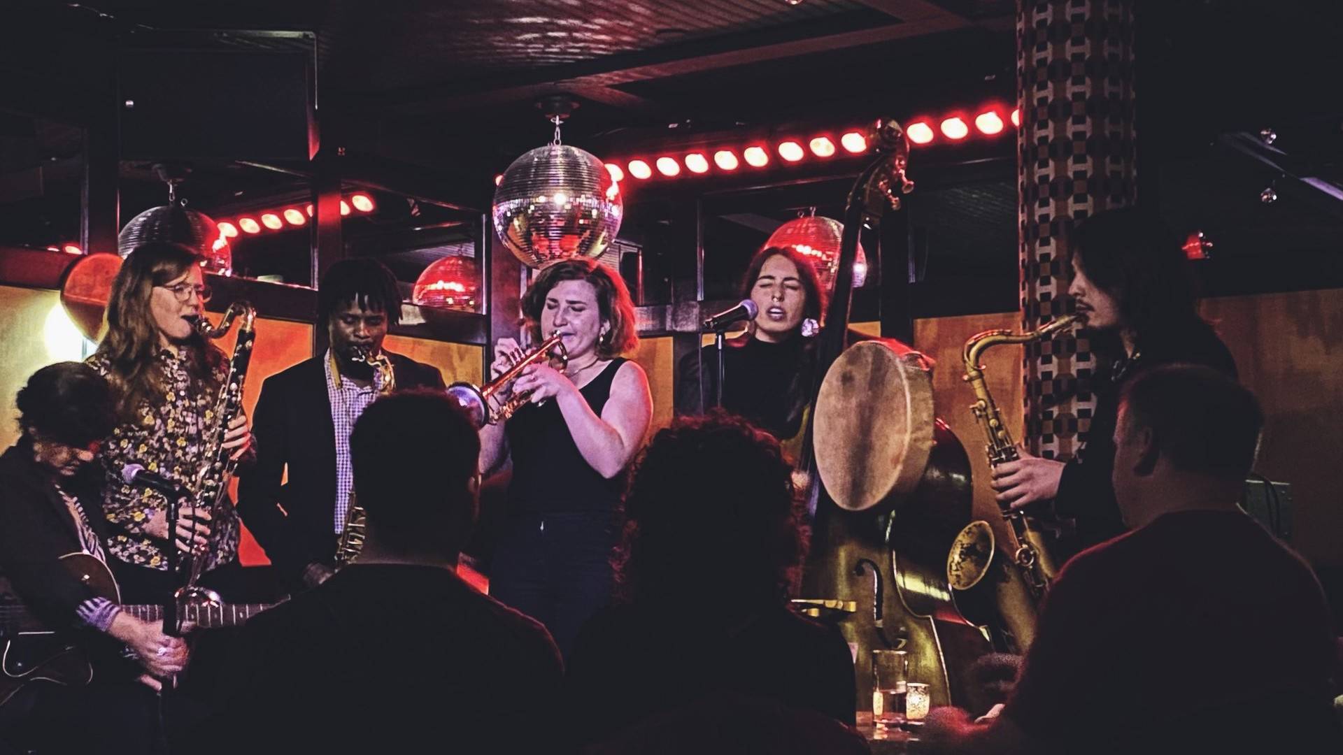 An band performing live. There is a guitarist, bass clarinet, trumpet player, bass player, and tenor sax player.