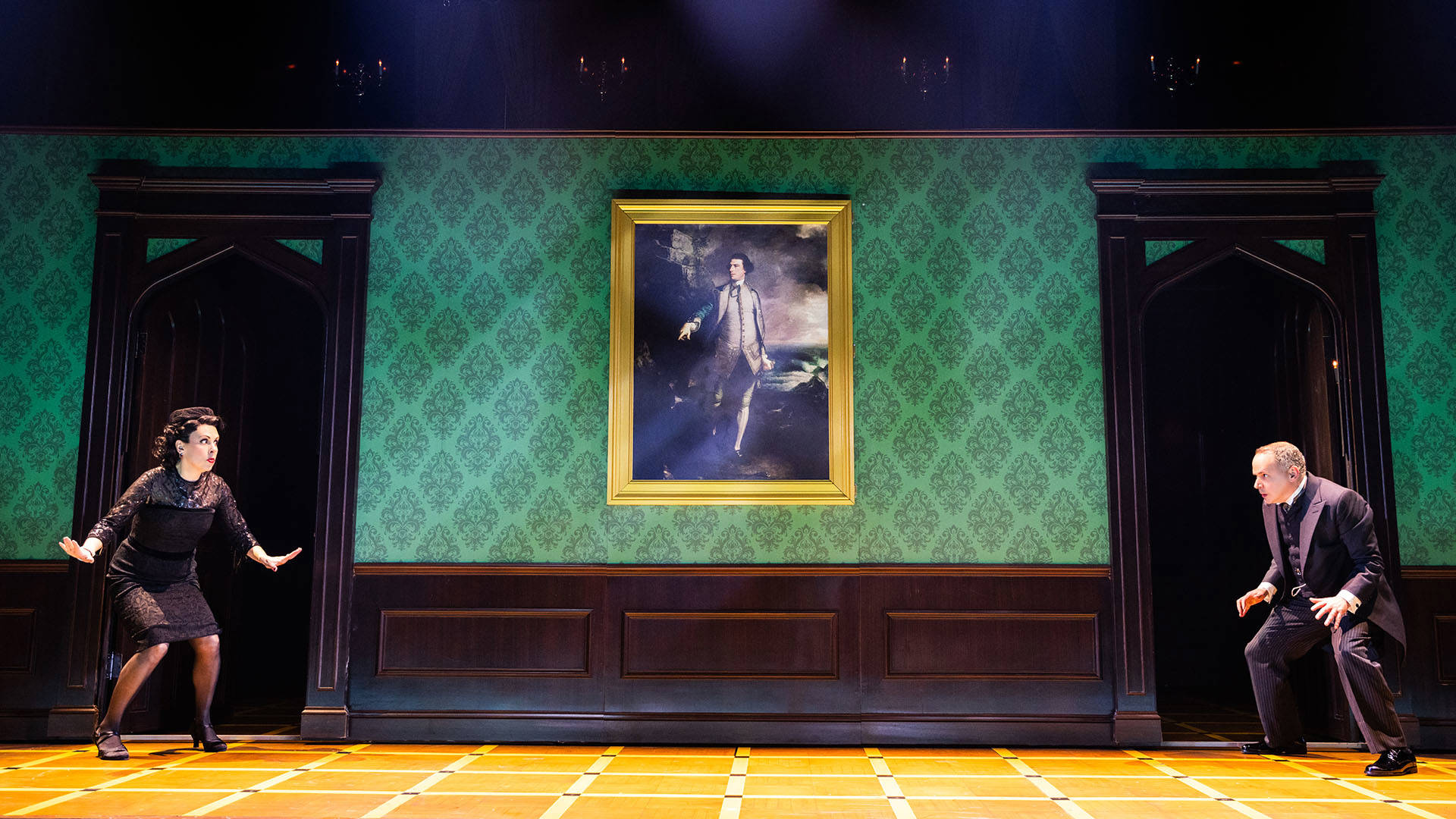 Two actors crouch in a standoff on stage, a long green hallway wall with a vintage portrait in between them.