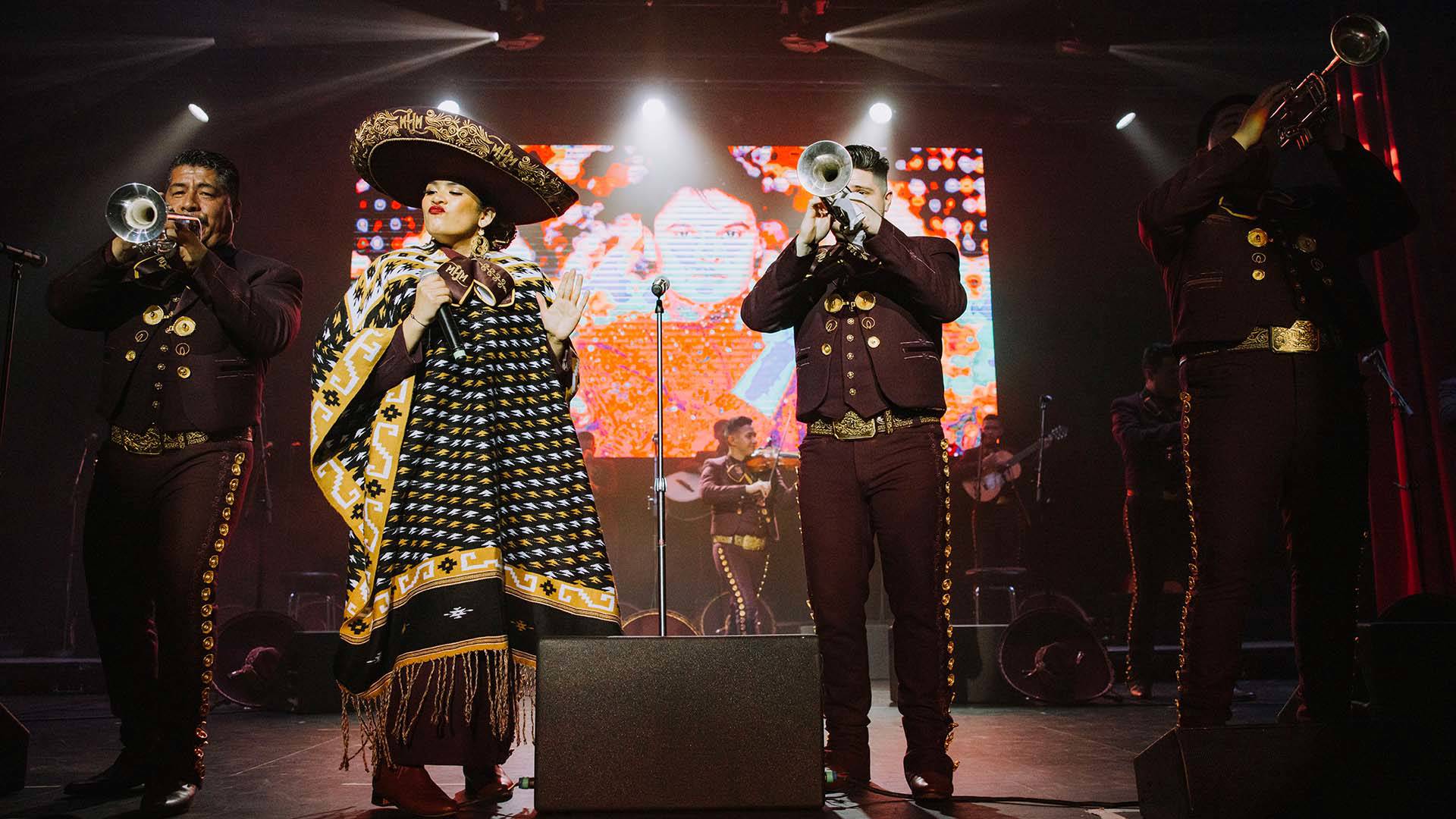 Four members of a Mariachi band standing at the front of a stage. Three are playing trumpets. They are wearing dark Mariachi outfits. The fourth is a singer wearing a sombrero and black and gold poncho. Behind them is a large screen with a woman wearing orange.