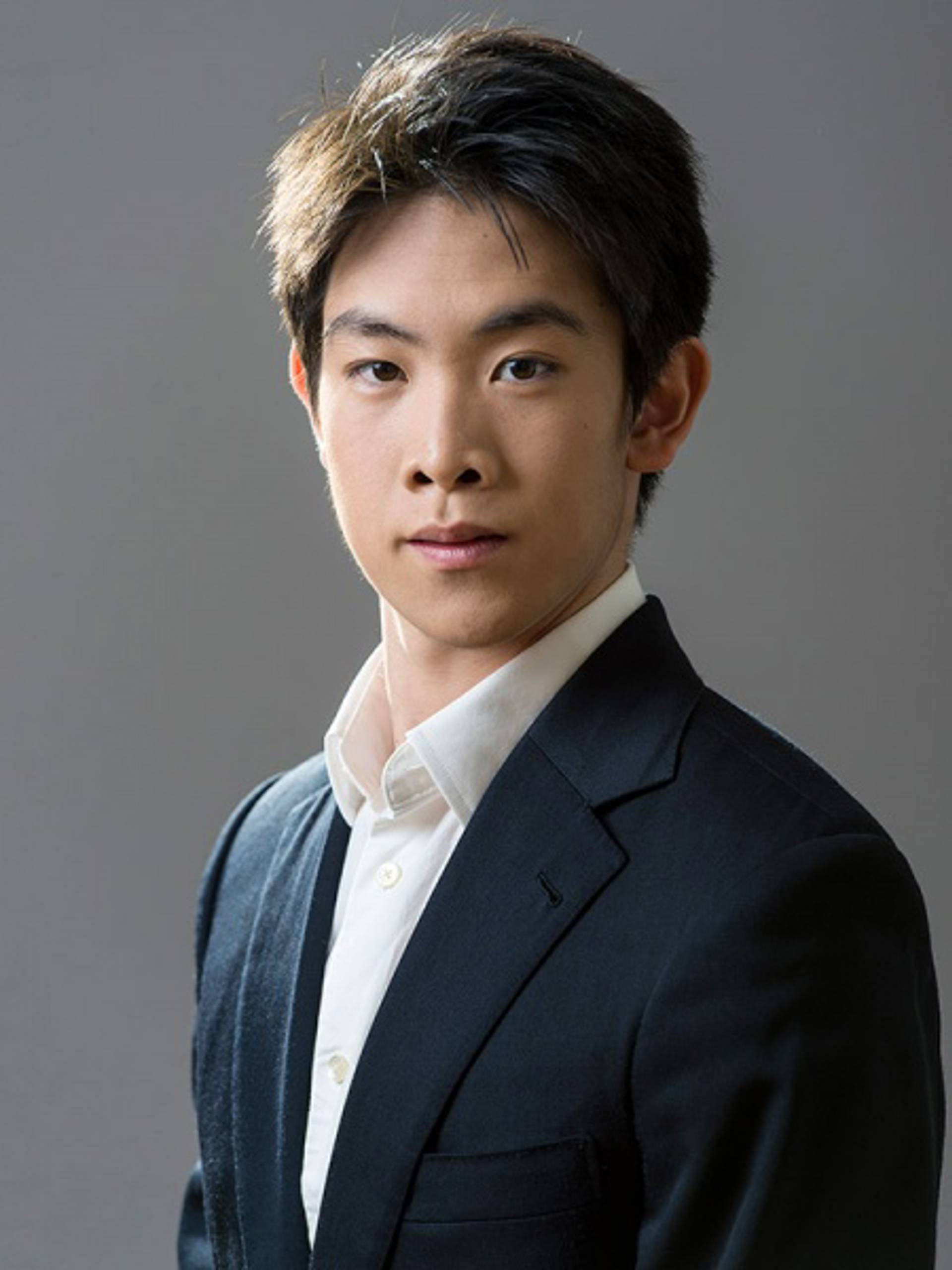 Headshot of a young man with medium complexion, short black hair, a white collared shirt and a dark jacket against a grey background