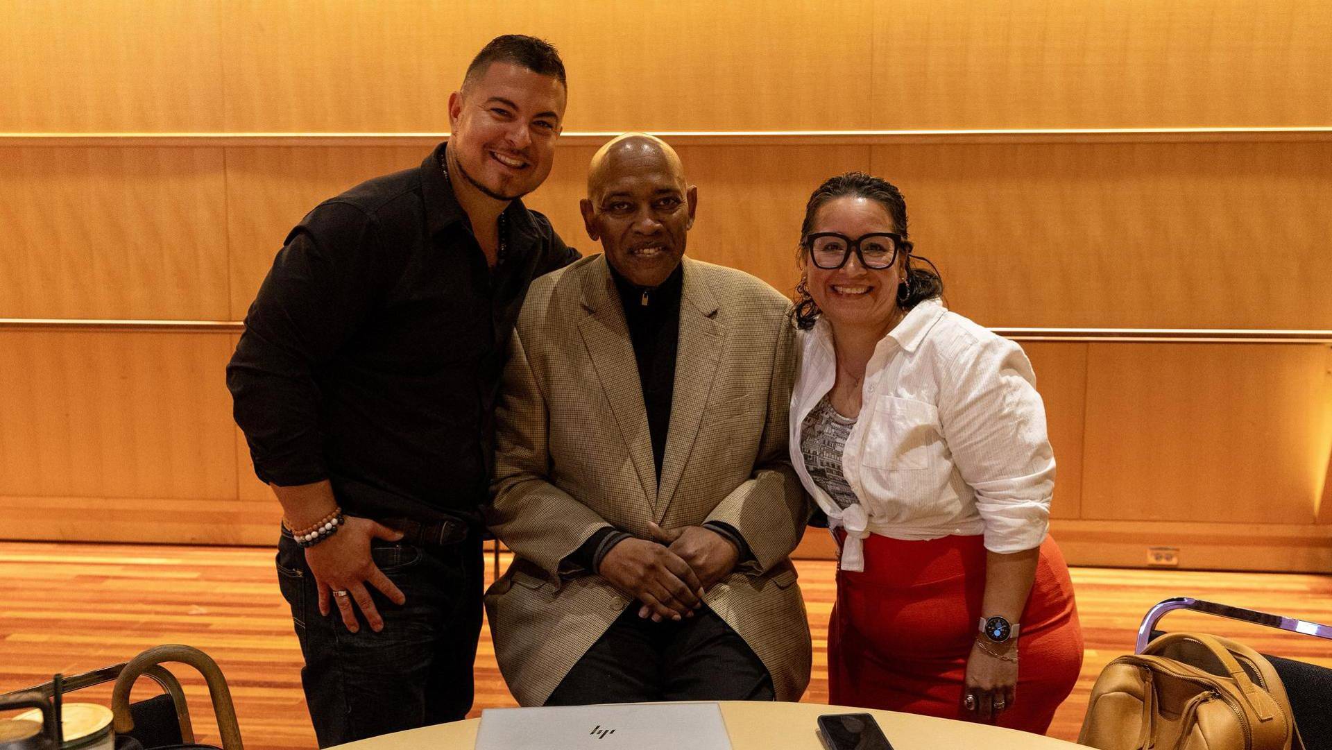 A man with medium skin tone, dark short hair and a black dress shirt poses with an older african american man with a bald head and tan jacket and a woman with medium complexion, dark hair, glasses and a white top.