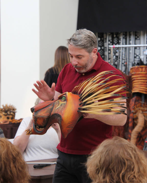 A man in a red shirt with grey hair holds a red wooden lion mask and is speaking about it.