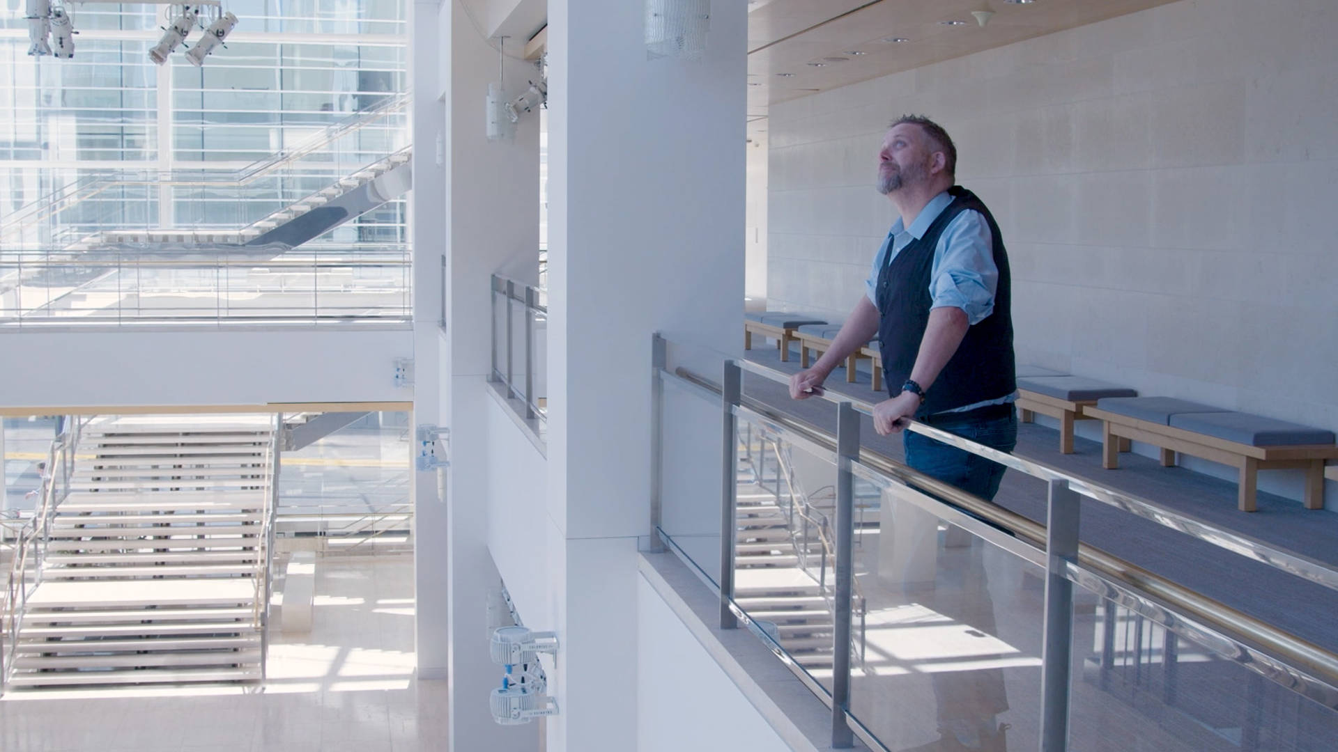 Jess Schuknecht standing against a railing in Overture Hall Lobby. He is a white man with short hai, a beard and blue eyes. He is wearing a blue shit and dark vest