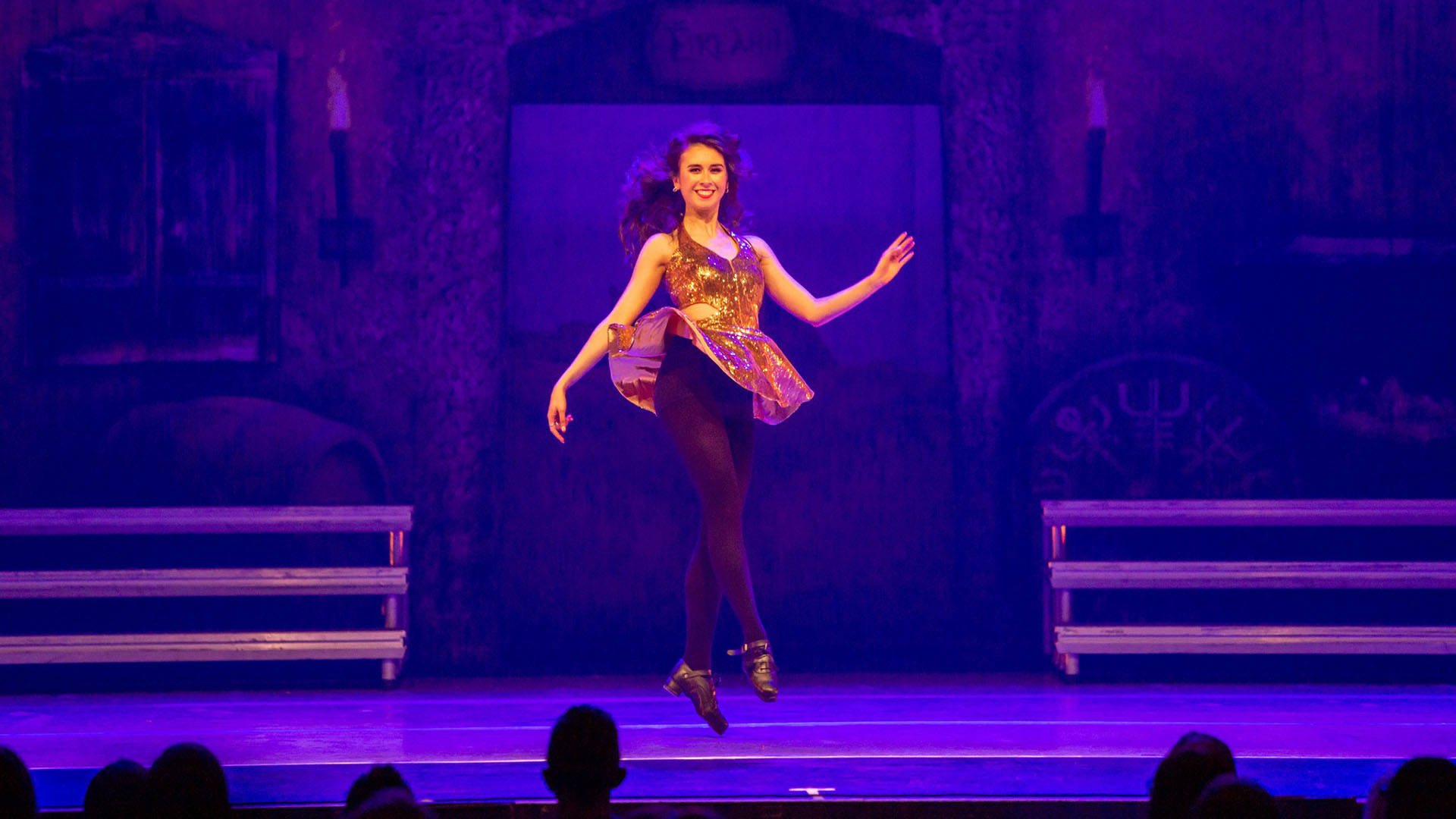 A woman in a gold dress with black tights performing irish dance on a purple stage.