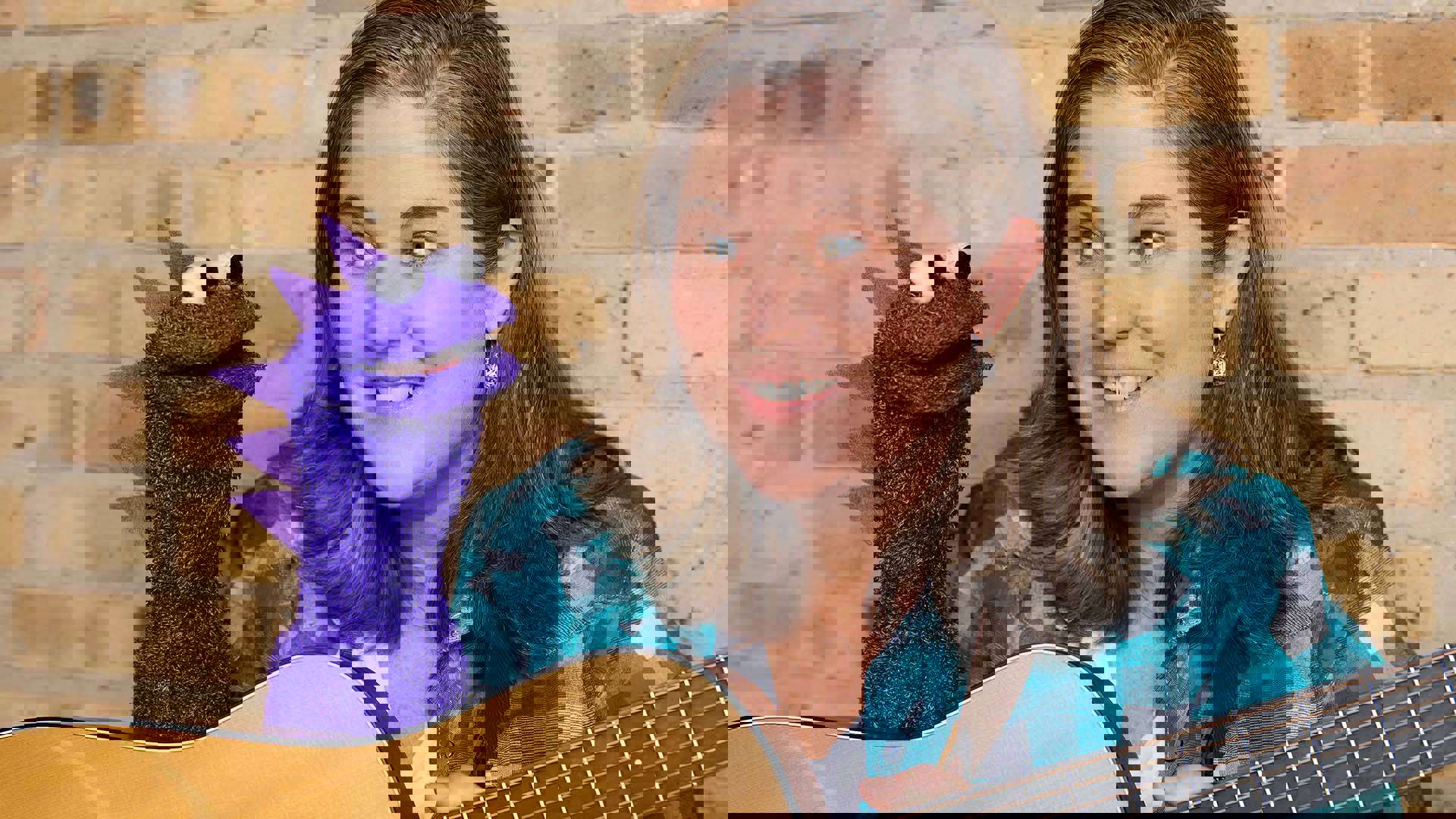 A white woman with brown hair holding a guitar. A purple, monster puppet is next to her.