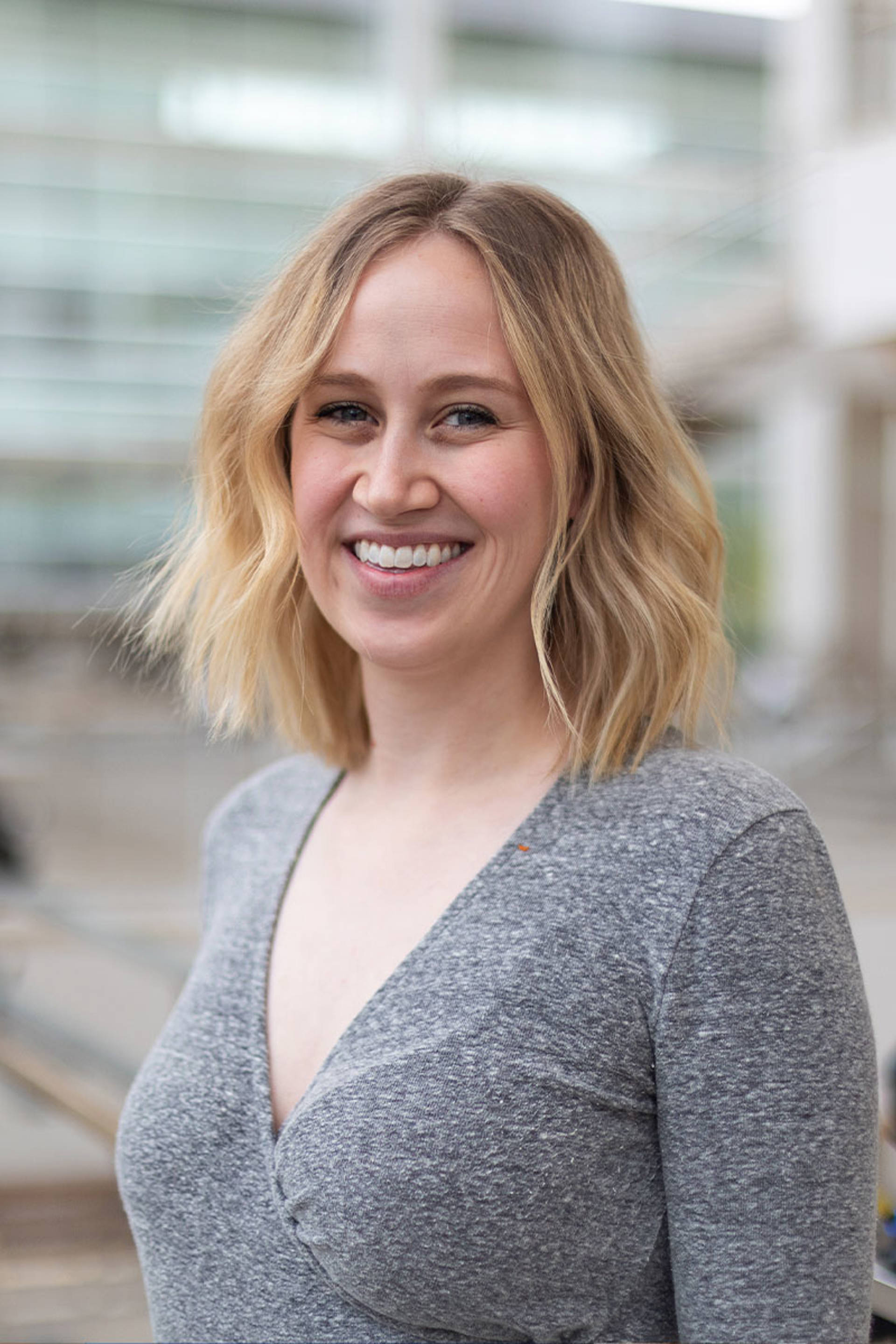 A white woman with blonde, shoulder-length hair and blue eyes. She is wearing a gray V-neck shirt and smiling.