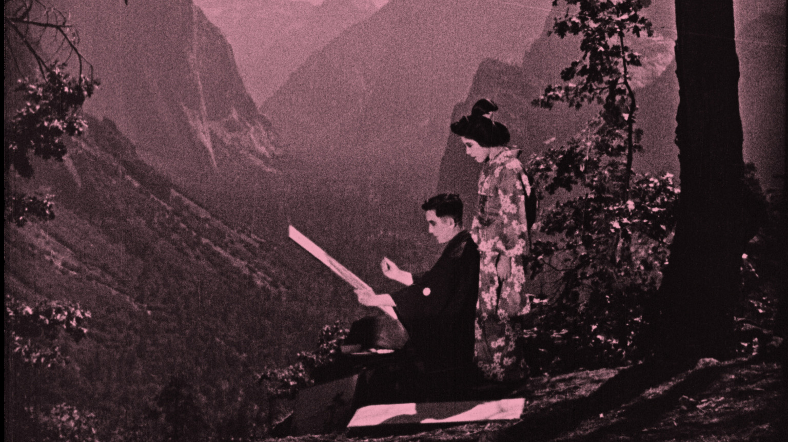 Black and White image of a seated man and standing woman with light complexion and dark hair. Both are wearing traditional Japanese outfits. The seated man is holding a canvas. They are outside with a mountain valley in the background.