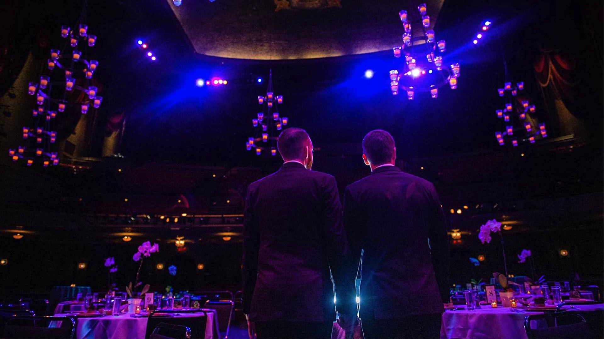 Two grooms standing on stage looking out into a theater. Chandeliers with candles hang from the ceiling.