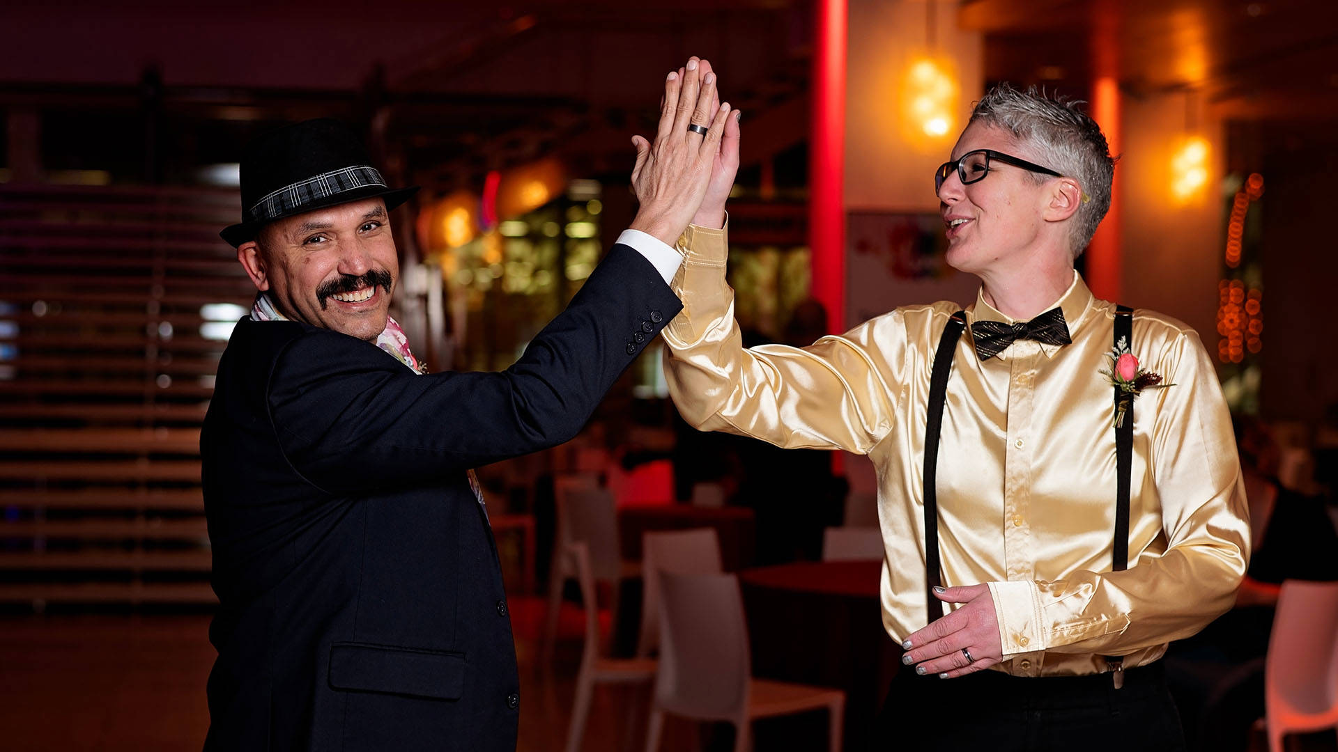 Two people high five. One is wearing a dark outfit with a fidora and a moustache. The other has short grey hair, a gold shirt and a bowtie.