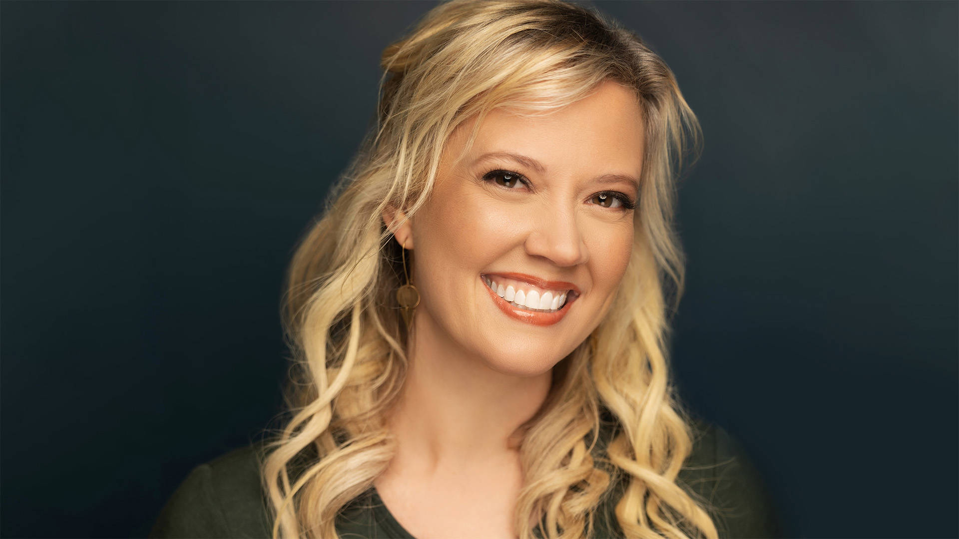 Headshot of Patti Murin, a white woman with long, curled blonde hair, smiling at the camera. She is wearing a dark green sweater against a black background.
