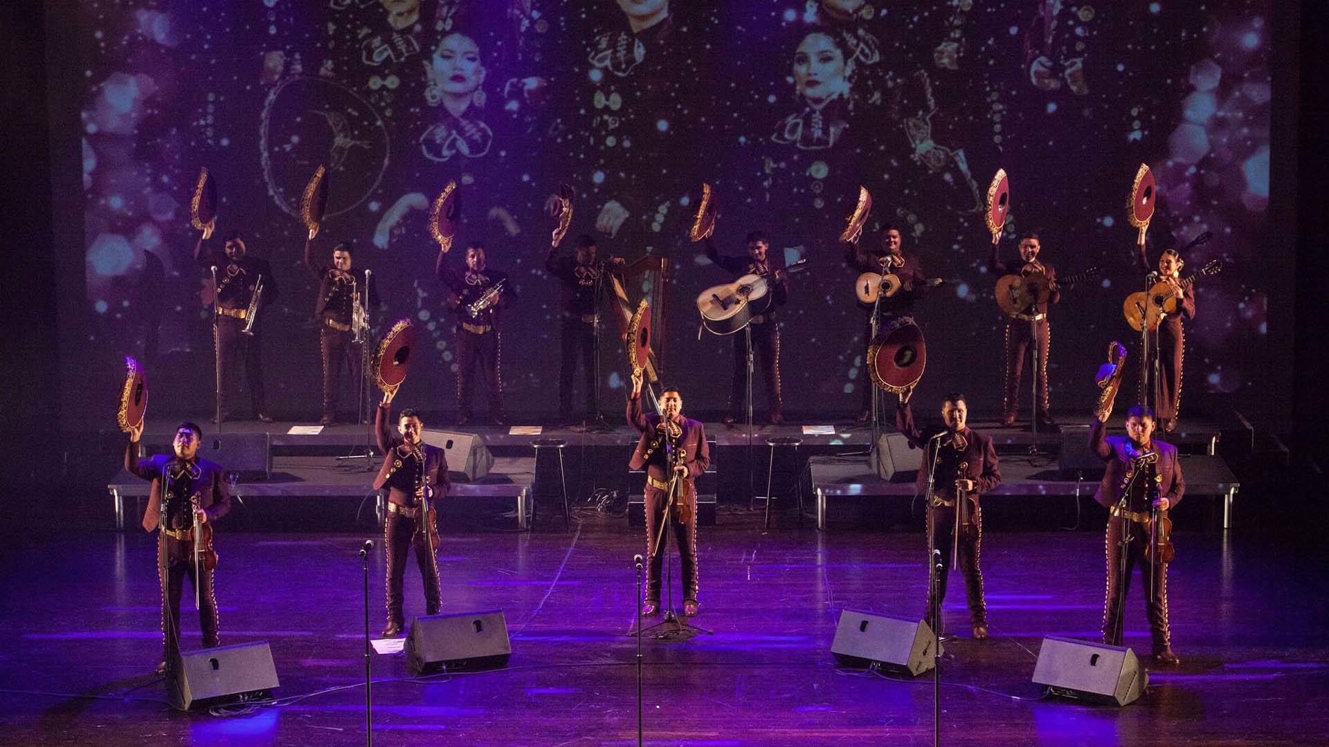 Two rows of people standing on a stage. There are five in the front row and eight on risers in the back row. They are wearing dark Mariachi outfits and holding sombreros in their right hands over their heads. They are holding violins, guitars and trumpets in their other hand.