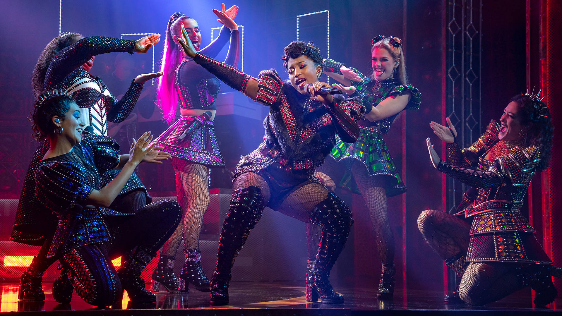 The cast of Six, in futuristic glittery outfits and holding microphones, performing on stage