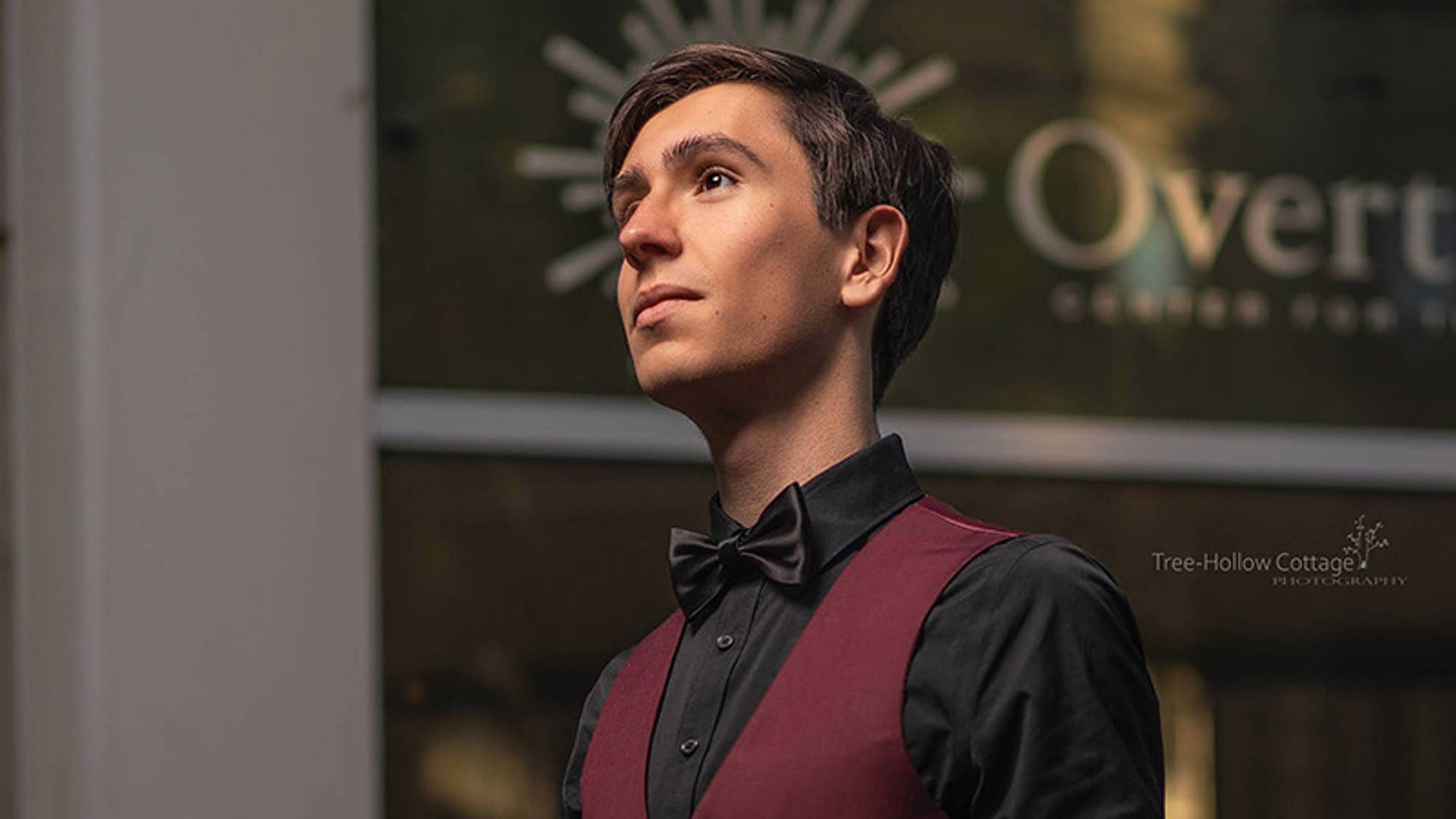 A caucasian high school aged boy posing in slight profile for a photo. He has short dark hair and a black button up shirt, crimson vest and black bowtie on.