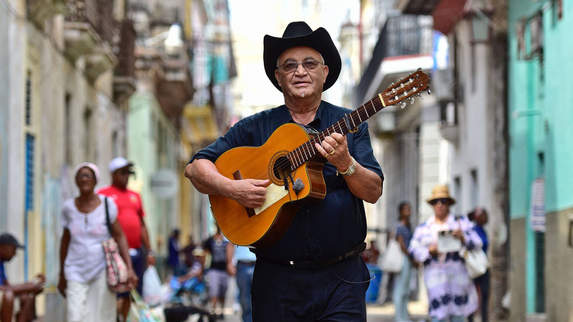 Eliades Ochoa, a Cuban man with medium skin tone and short white hair. He is wearing tinted glasses, a black cowboy hat and a black dress shirt while playing the guitar in a street.