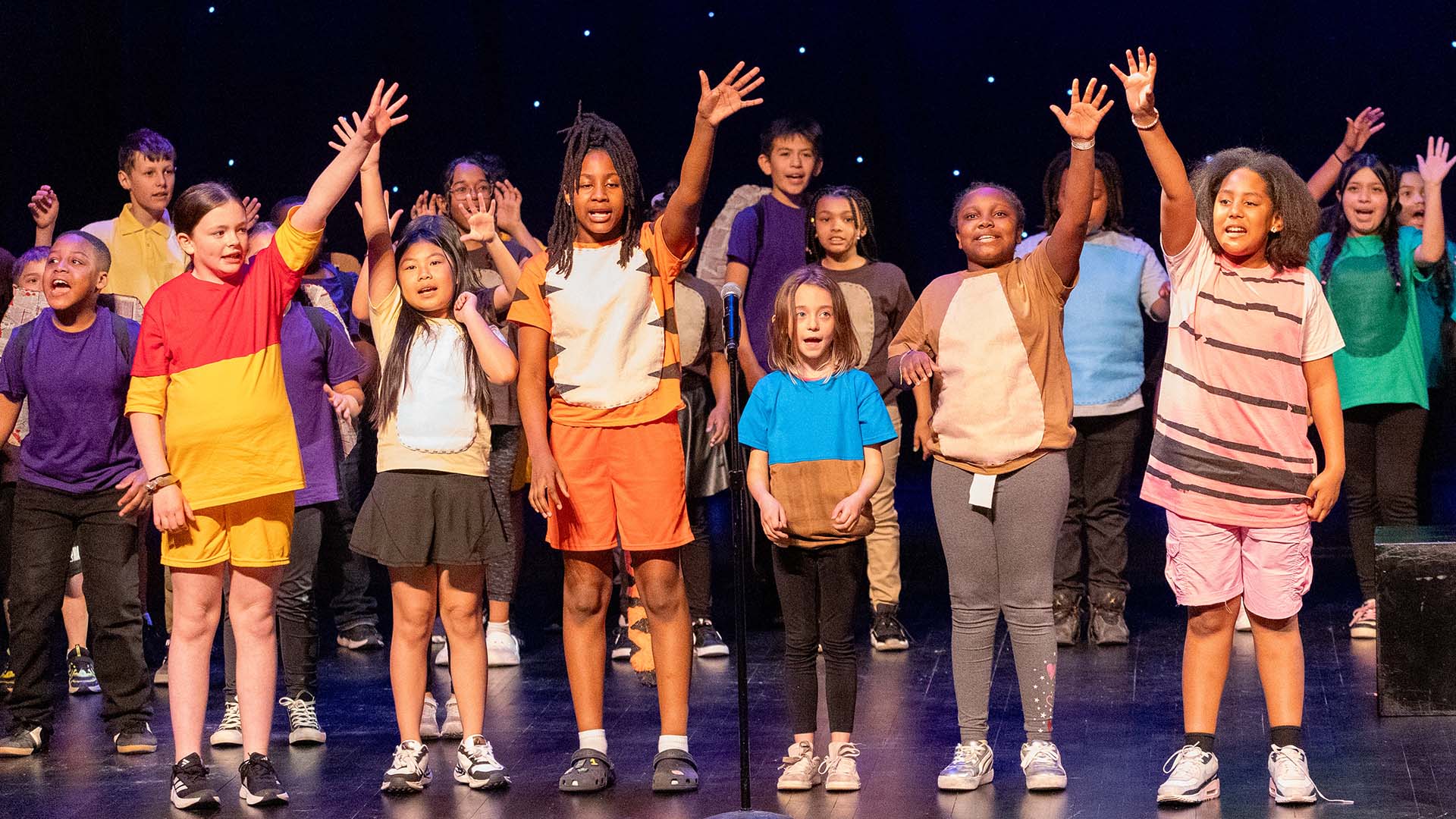 Young students in colorful costumes performing on a dark stage. 