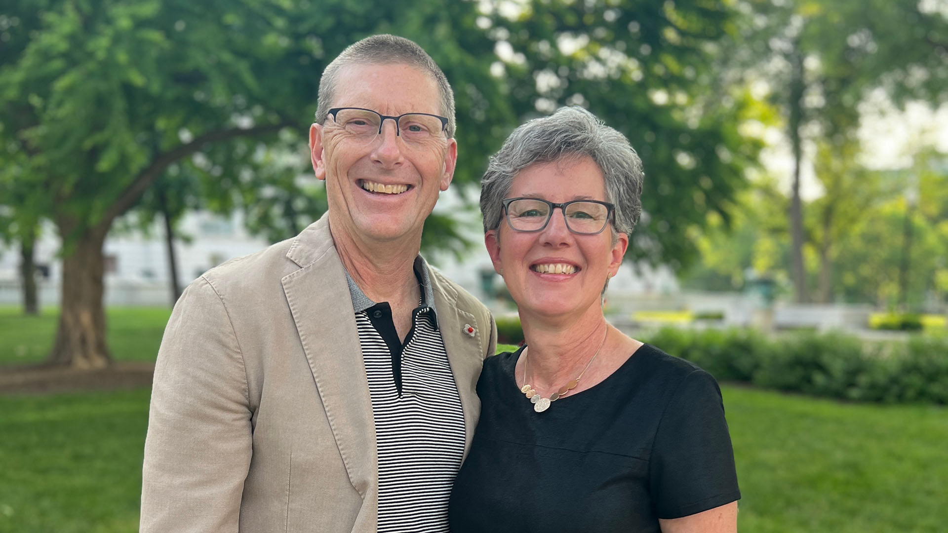 Kevin and Lorrie Meyer, a middle-aged caucasian couple smiling together outside. One has a buzzcut with glasses, wearing a tan blazer and polo shirt. The other has short grey hair with glasses, wearing a gold necklace and black blouse.