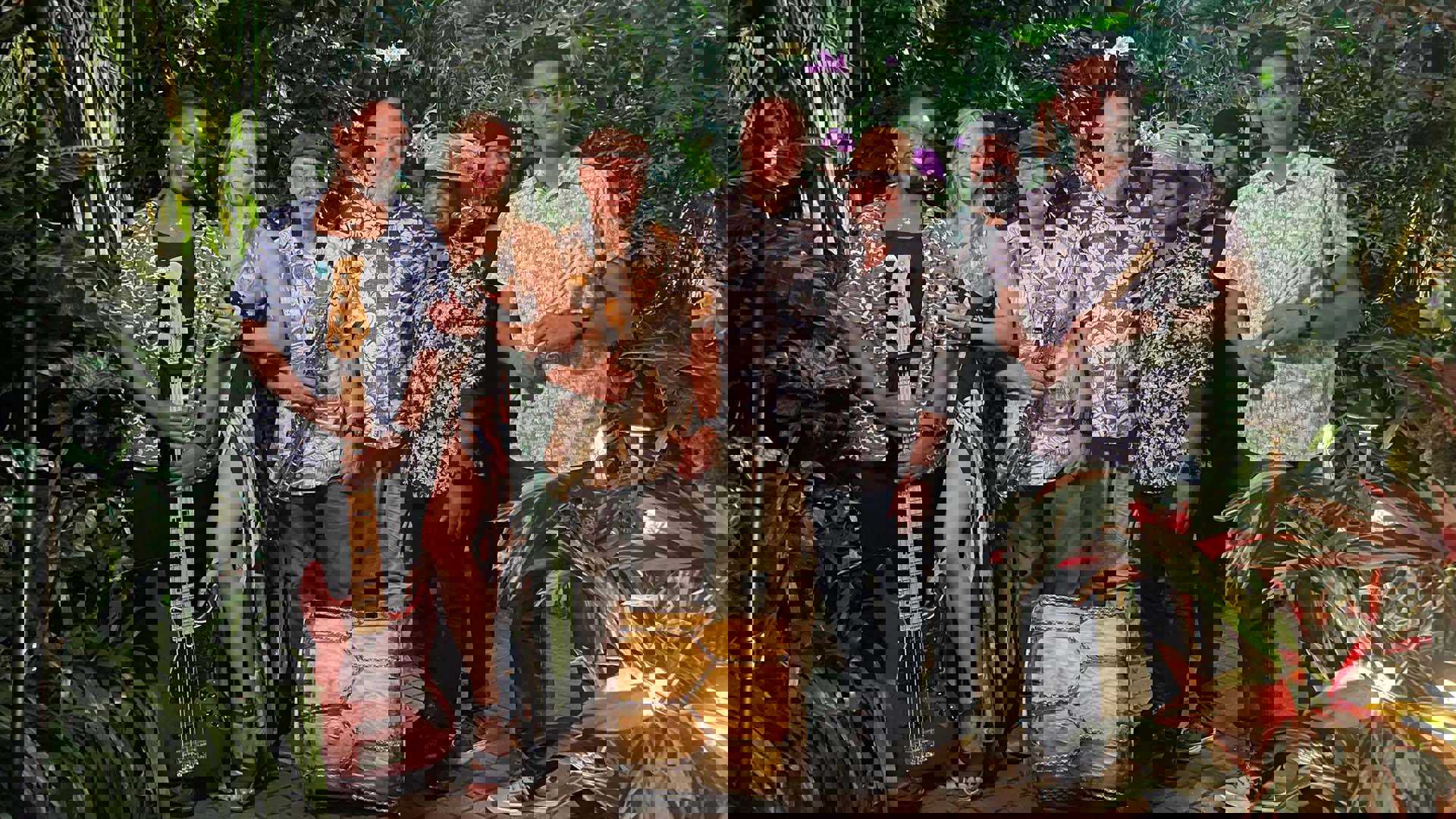 Seven members of a band in colorful shirts posing for a photo. They are surrounded by plants They are holidng several instruments - a bass guitar, soprano saxophone, and a drum.