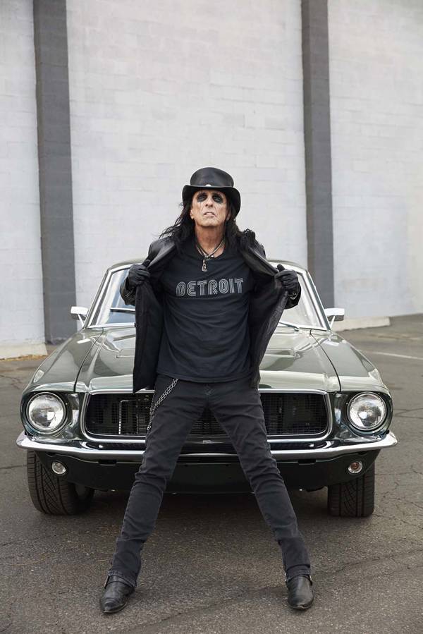 An older man with long, black hair and a lot of eye shadow. He is in black jeans, t-shirt, leather vest and a top hat and is leaning against the hood of a car. His t-shirt reads "Detroit".