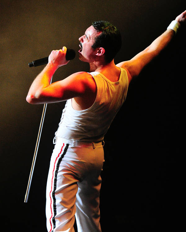 A caucasian man with short black hair and a moustache singing into a microphone with his right hand outstretched. He is wearing all white with a tank top.