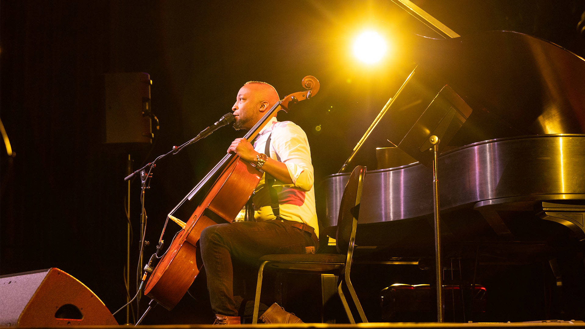 An African American man with a short mohawk and beard sitting and singing into a microphone. He is holding a cello in playing position and there is a grand piano behind him.