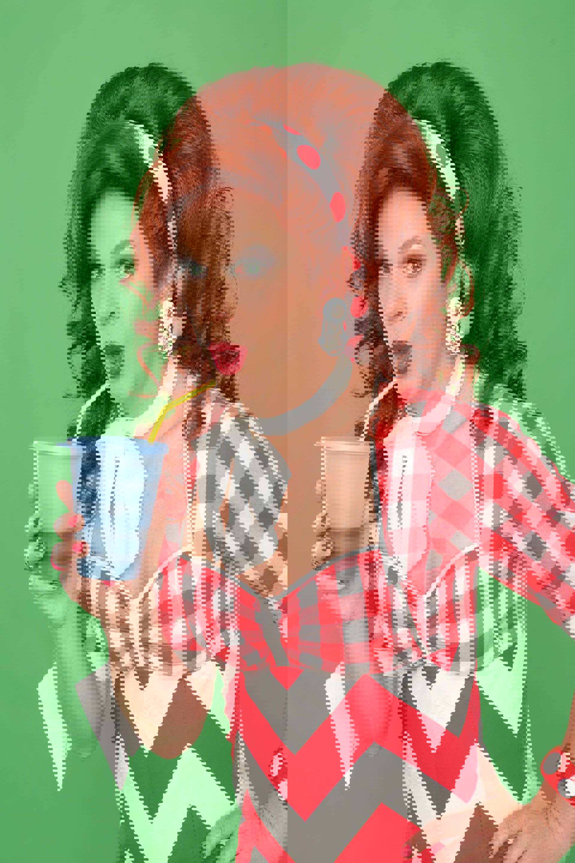 A drag performer with voluminous red hair sipping from a blue cup and yellow straw in a red and white gingham dress and headband.