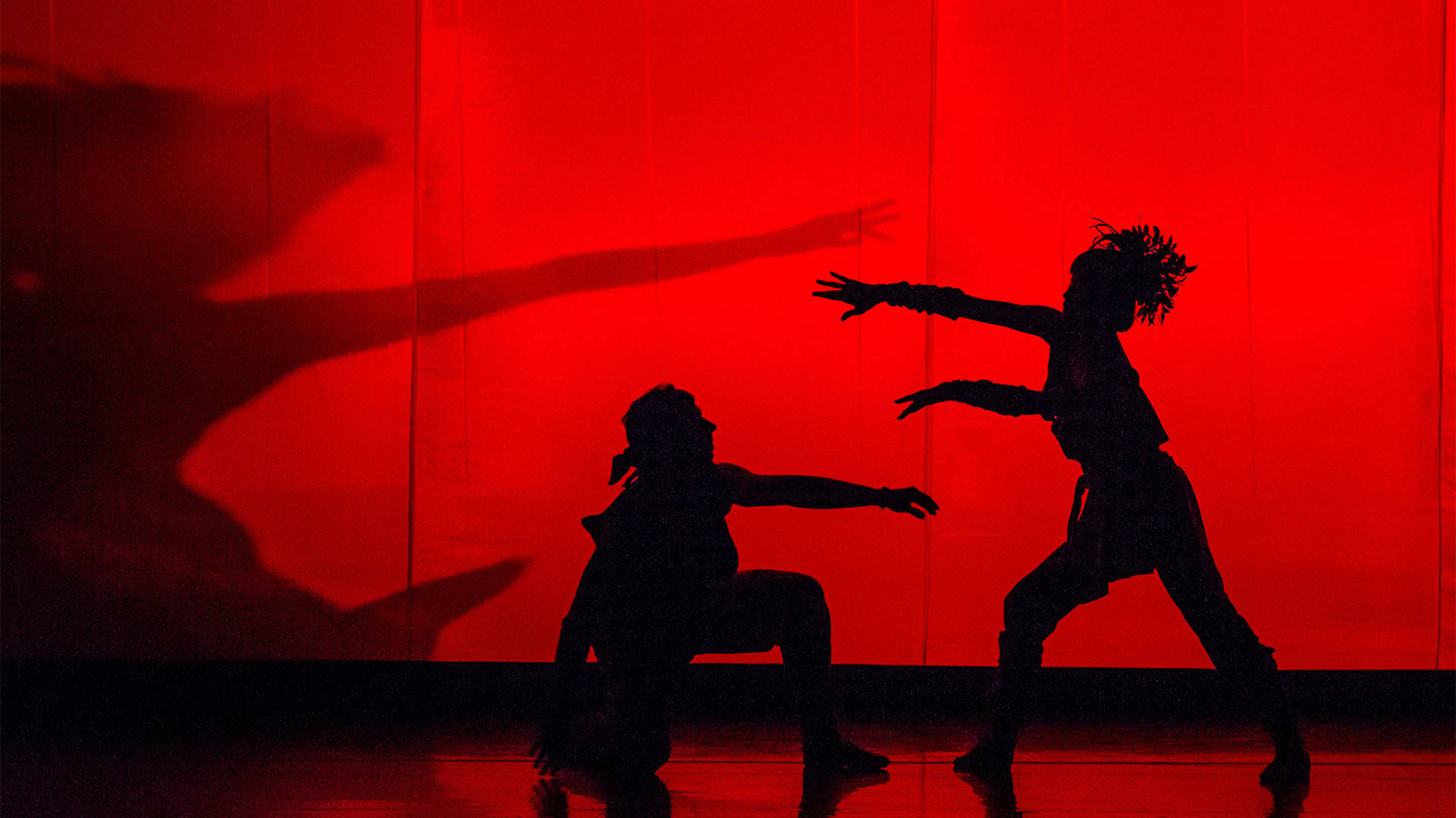 Two dancer silhouettes performing in loose clothing against a deep red lighting backdrop.