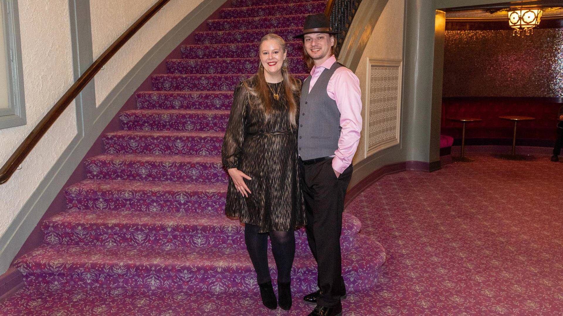 A man and a woman pose for a picture in Capitol Theater Lobby. He is wearing a pink dress shirt with a grey vest and fedora. She is wearing a gold and black dress.