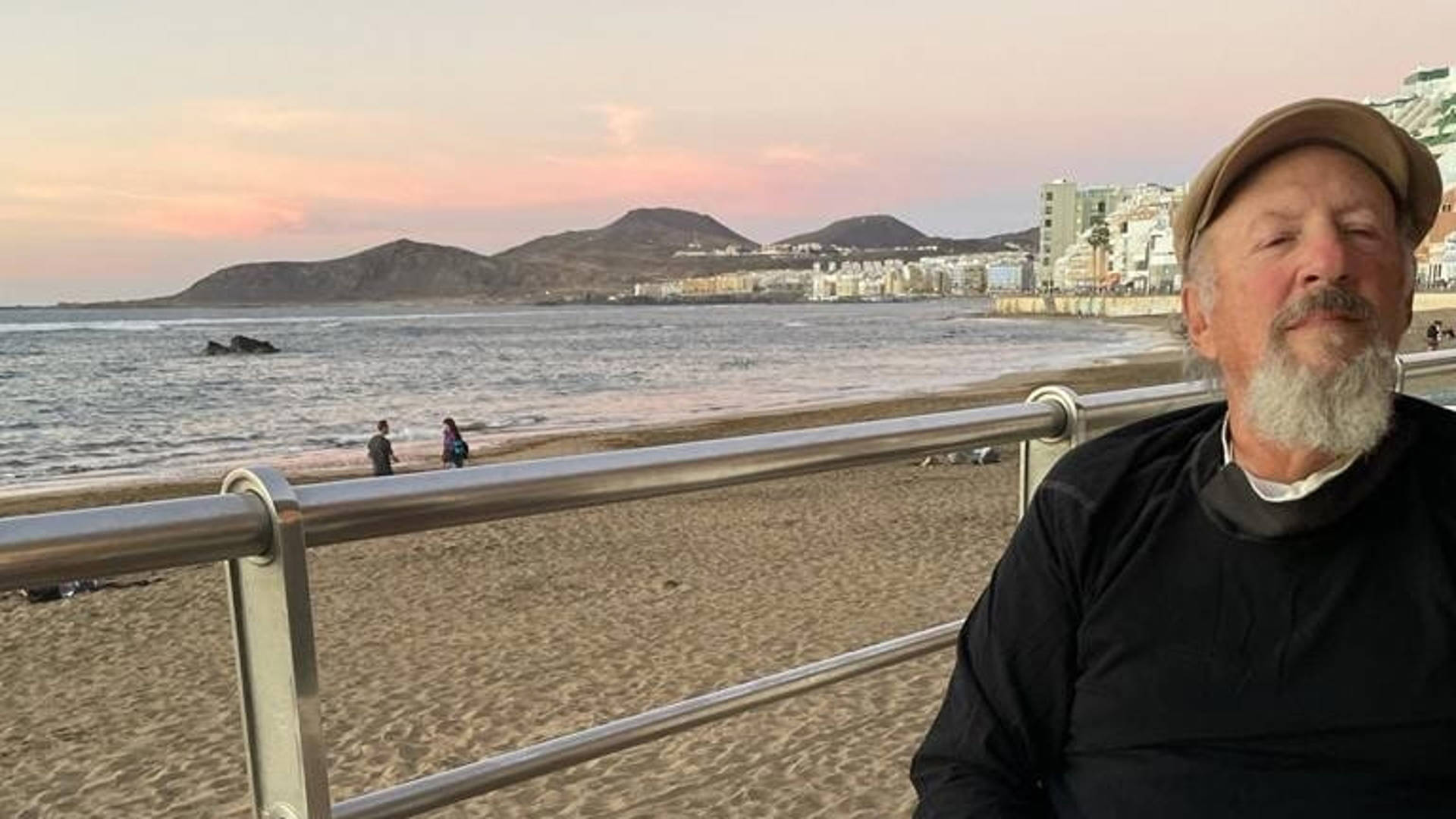 An older white man with a grey and white goatee in a cap sitting by an ocean and beach.