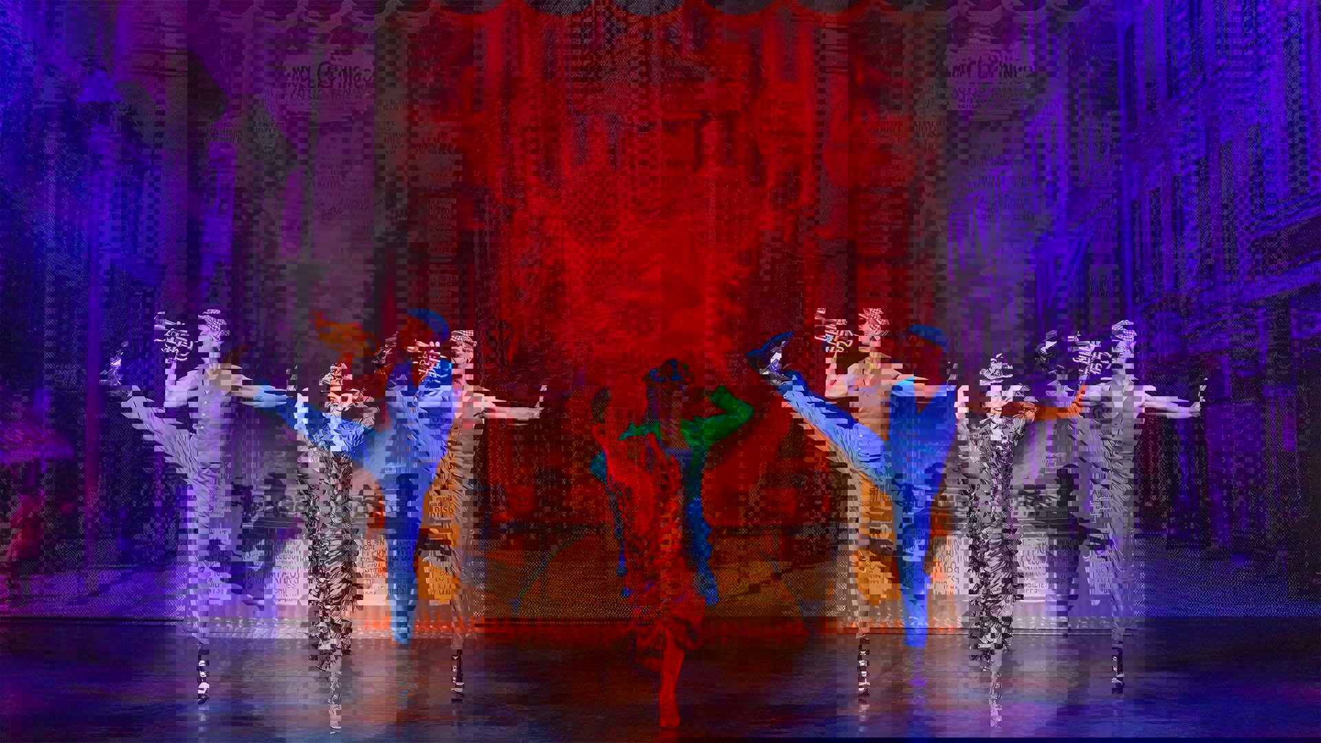 A light complexion woman in a colorful dress kicks her leg up high in the air, performing on stage. Two men in vests and playing trumpets kick up behind her in a line.