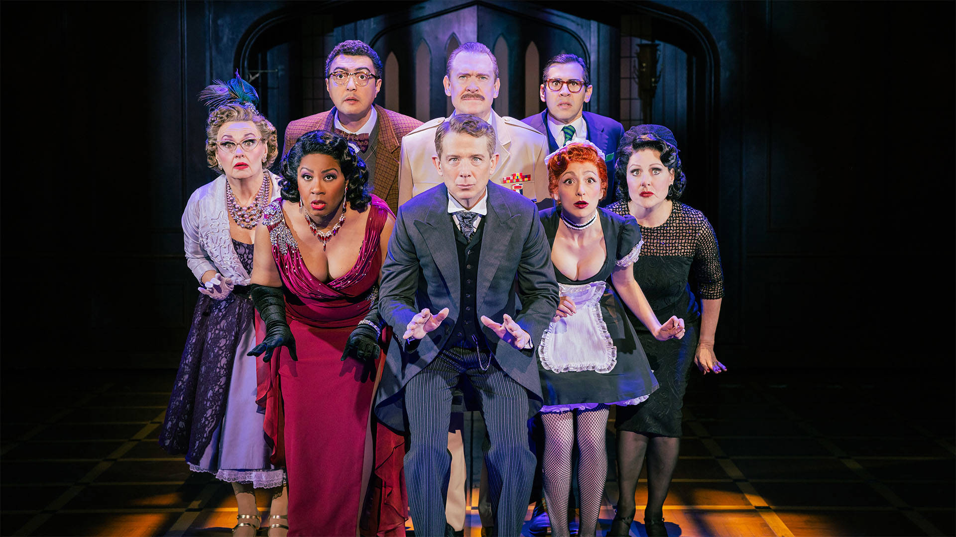 Eight Clue actors huddled together looking with concerned faces to their left. Three of the women are wearing formal dresses and fourth is wearing a black maid outfit. The four men are wearing various suits.