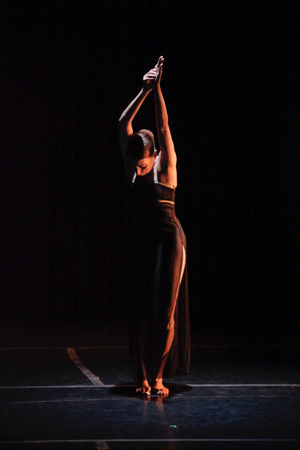 A woman on a stage with dark surroundings in a dance pose with her hands held together above her head. She has light complexion, dark hair and a sheer dark outfit on.
