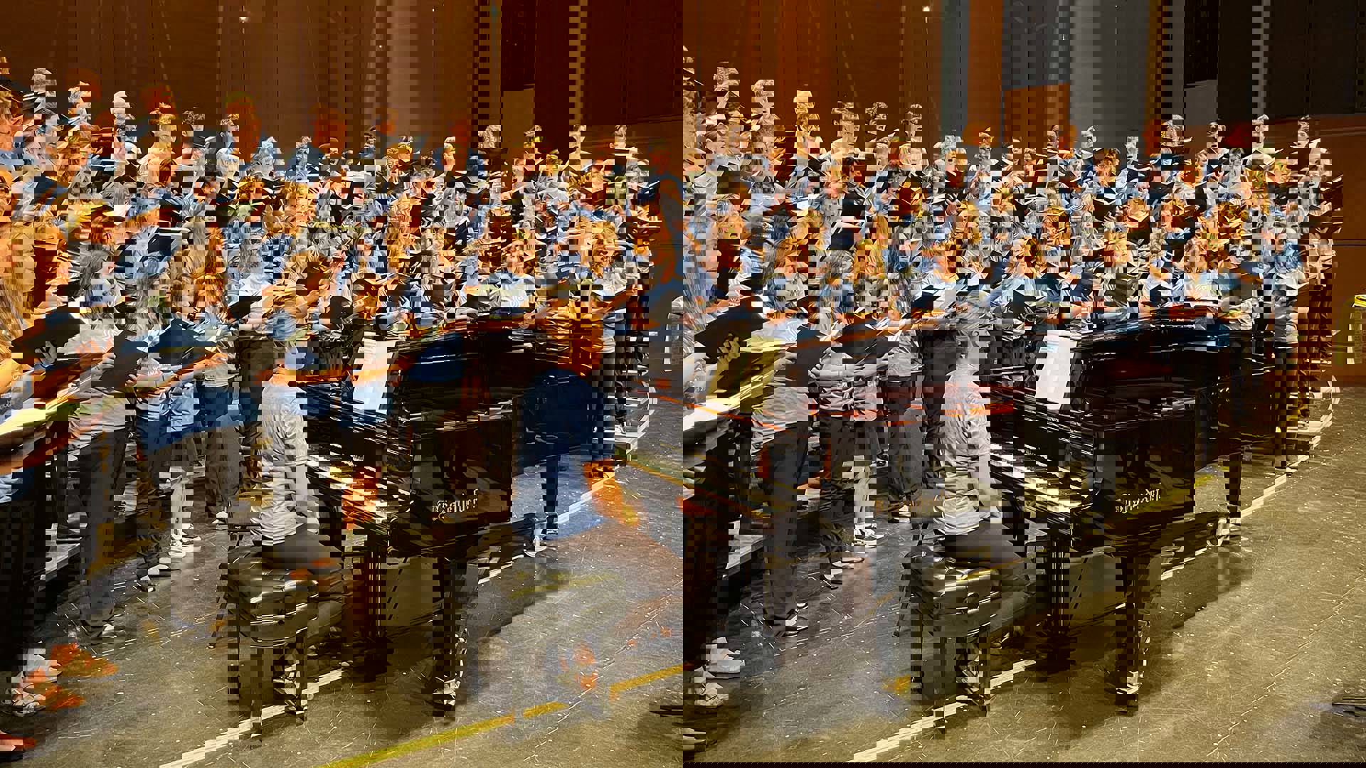 A large choir of high school students in blue t-shirts and black pants practicing on stage with a grand piano.