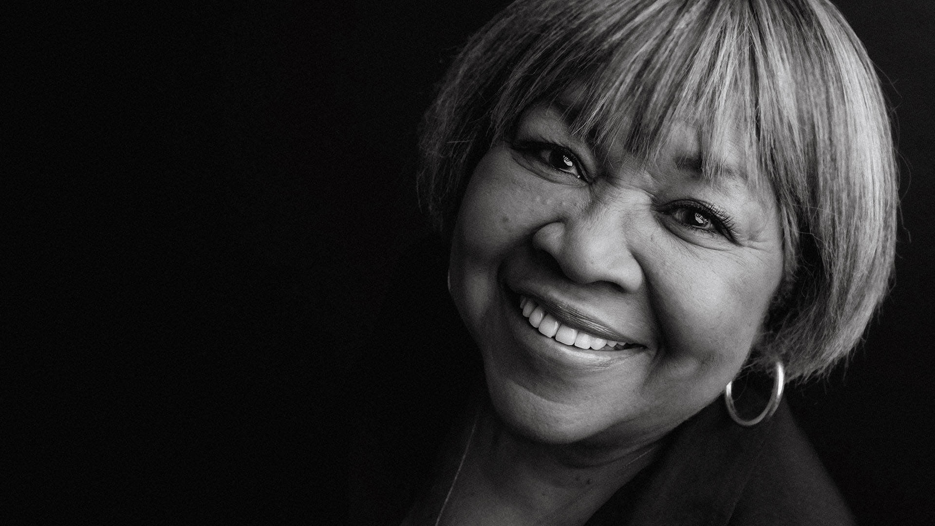 Black and white image of Mavis Staples. She has medium length hair and is smiling warmly to the viewer. Dark background