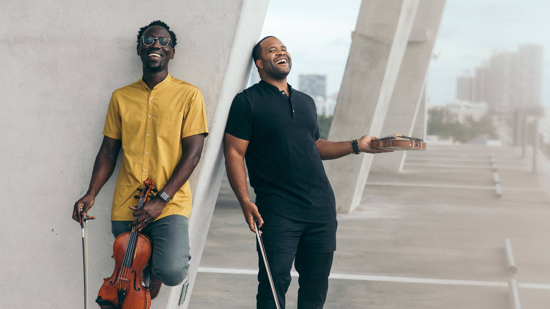 Two laughing men holding violins and leaning against a cement column. The man on the left has dark complexion, glasses and a yellow shirt. The man on the right has dark complexion and a dark shirt.