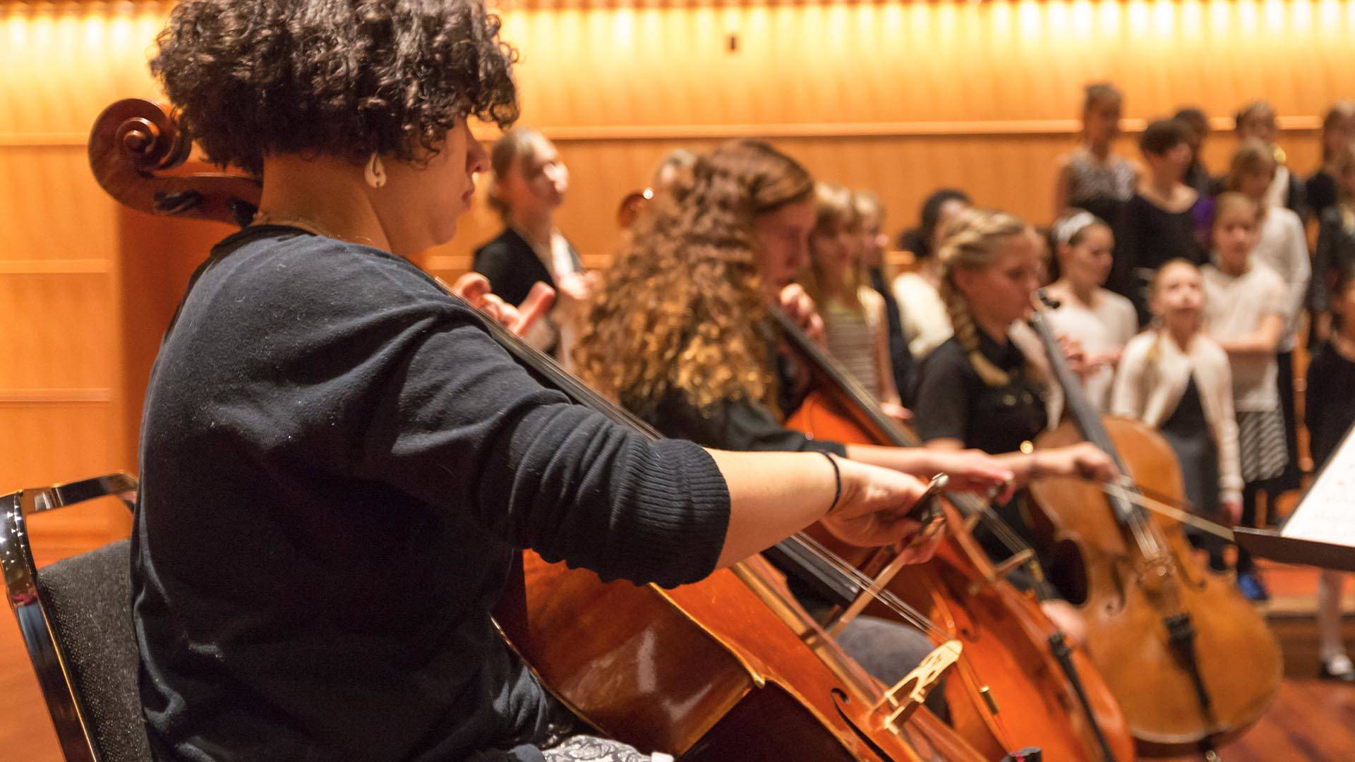 A person with curly dark hair playing a cello. There are other cellists out of focus next to her.