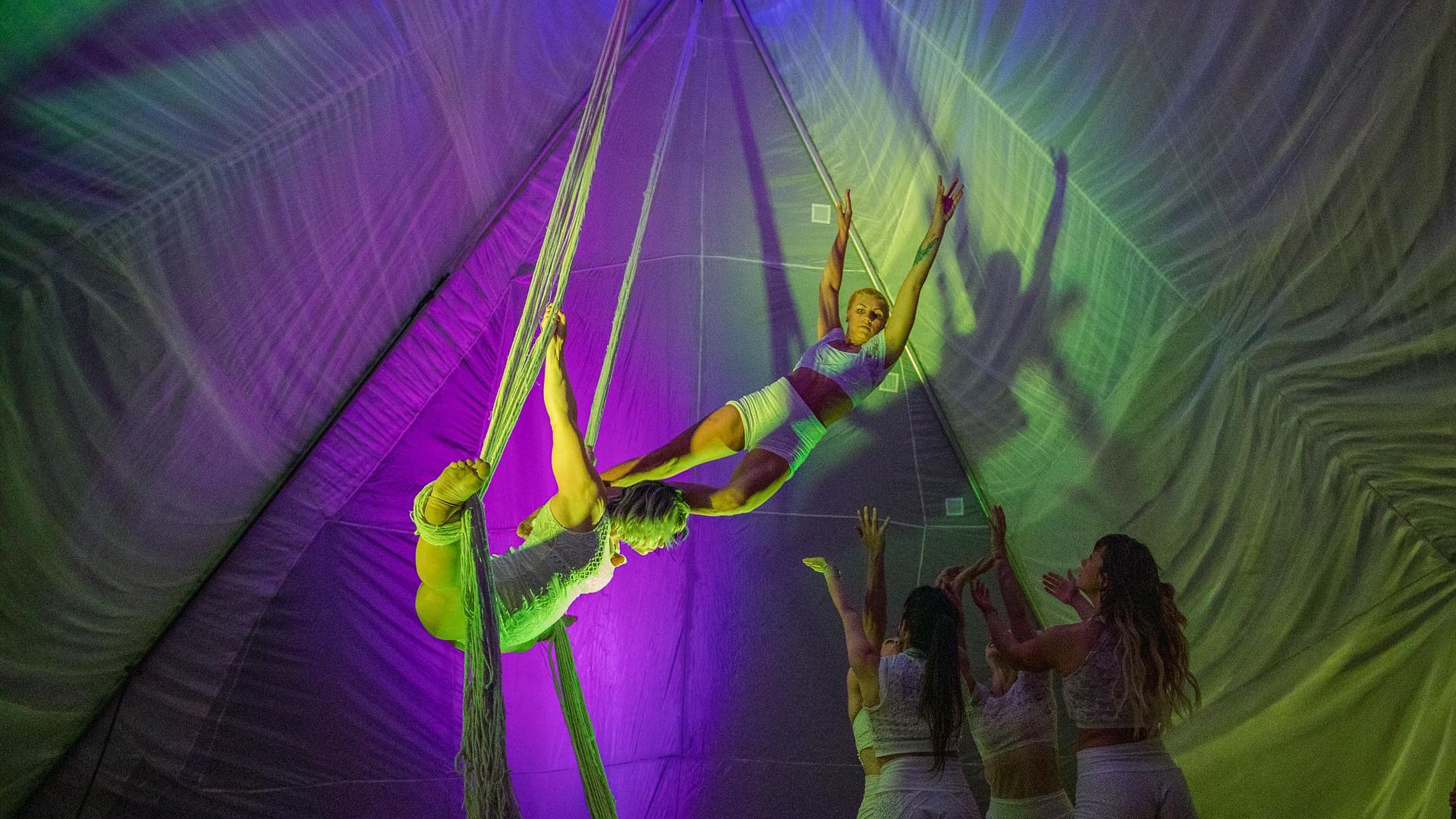 Two acrobats with light complexion are spinning together from two suspended white ropes. A group of acrobats stand below them to spot. All of them are in a large white nylon tent with purple and green lighting.