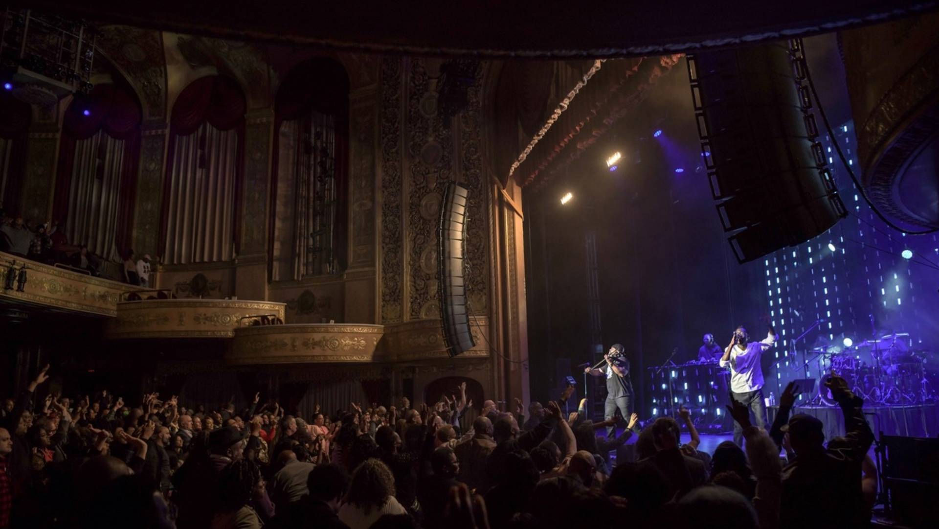 Wide shot of Black Violin performing to an animated crow in a theater. On stage you can see one man playing a violin and the other singing into a microphone.