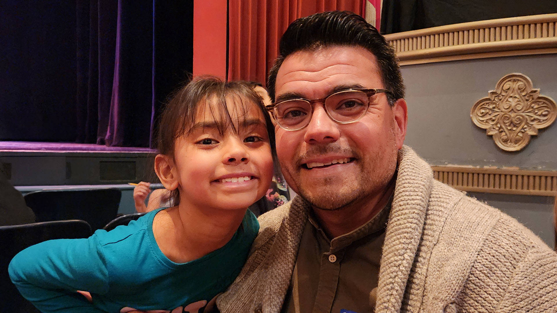 Two people smile for a photo together in Capitol Theater. On the left is a young girl with medium complexion, brown hair, and brown eyes wearing a blue shirt. On the right is a middle aged man with glasses, dark hair and beige knit cardigan and buttondown shirt.