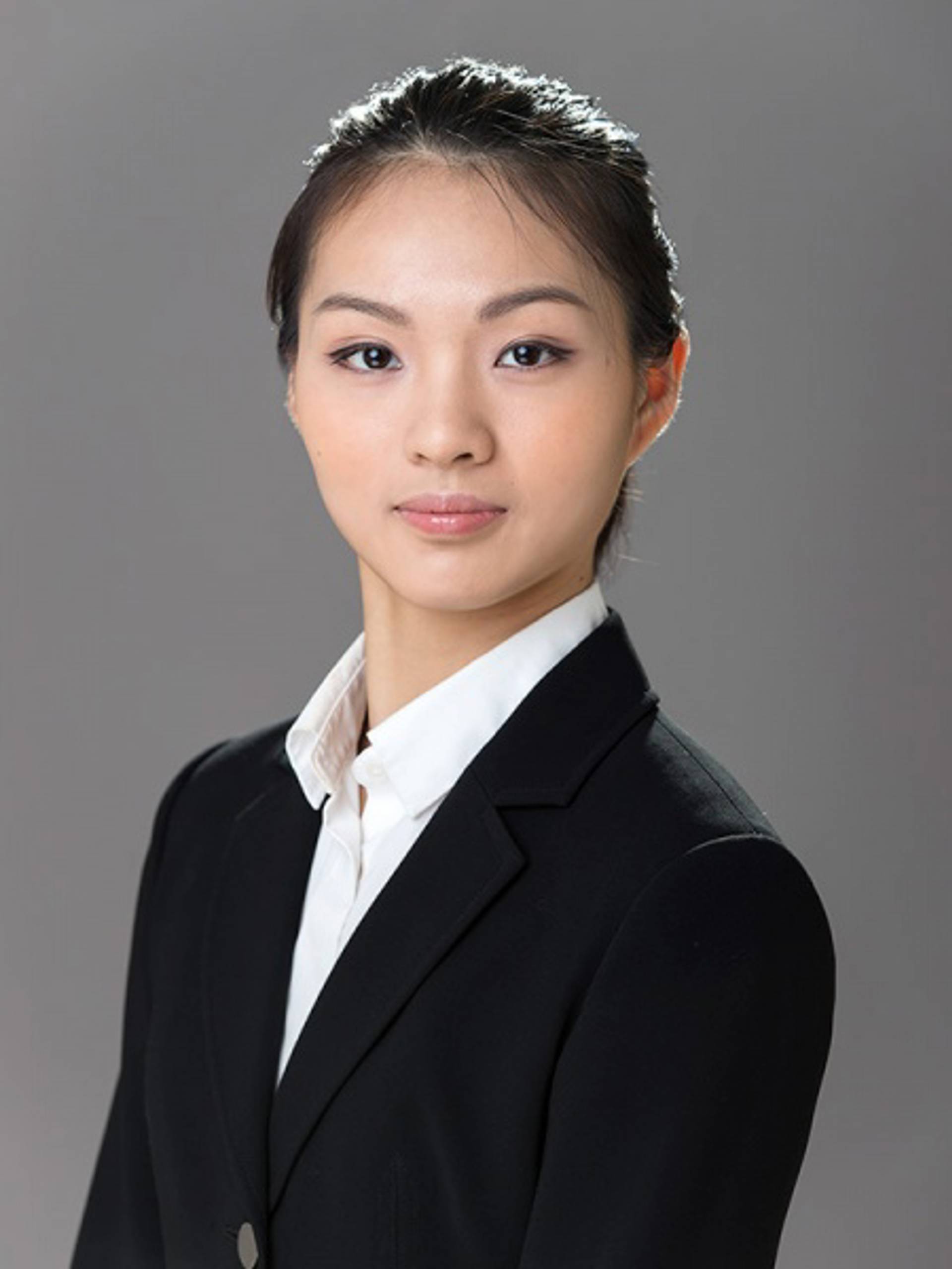 Headshot of a young woman with medium complexion, long black hair, a white collared shirt and a dark jacket against a grey background