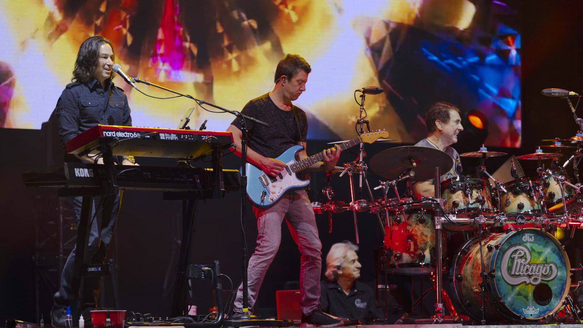 Three men on stage playing a keyboard, guitar and drumset.