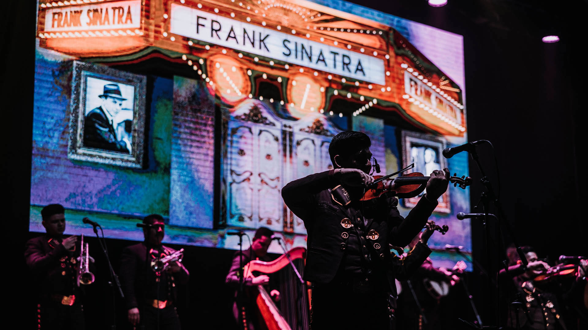 A silhouette of a violin player. They are wearing a dark Mariachi outfit. Behind them is a screen with an image of a building. The building has a marquee that reads "Frank Sinatra". Below the screen are additional Mariachi players holding trumpets and string instruments.