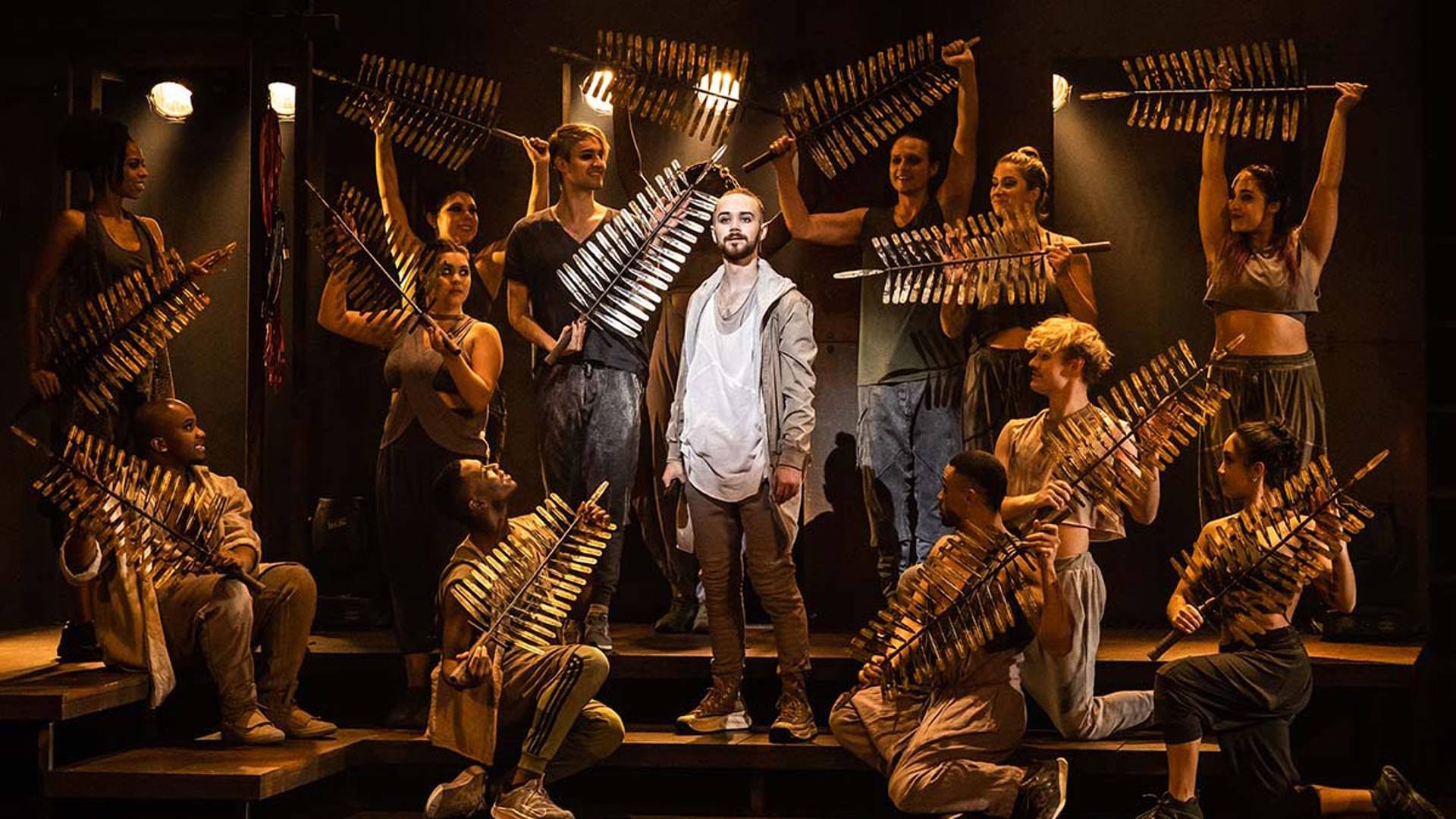 A caucasian man stands in the spotlight with the ensemble surrounding him holding golden palm leaves.