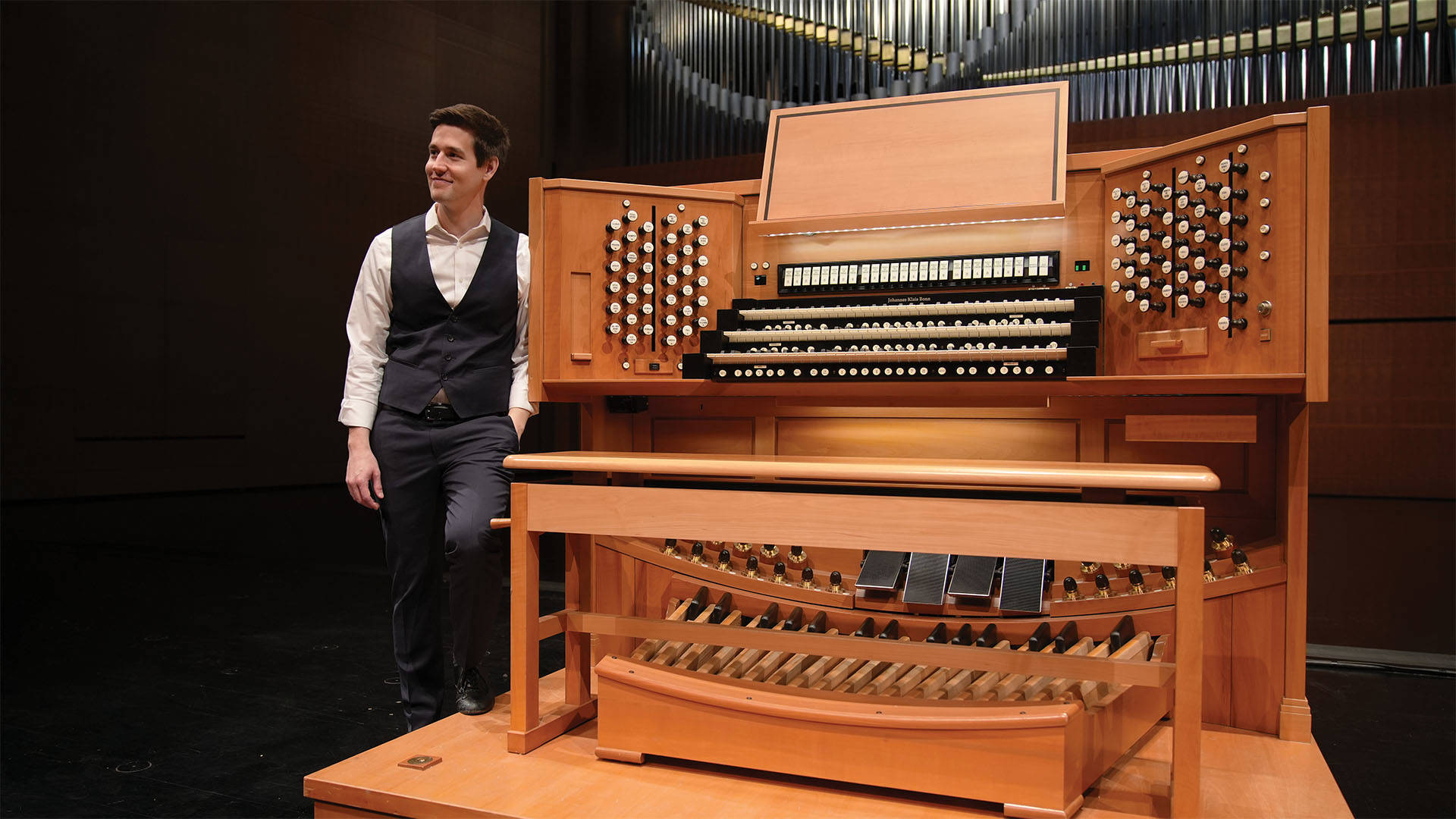 Greg Zelek, a young man with light complexion, short styled brown hair, a white button-up shirt and black vest, standing next to a large wooden organ console.